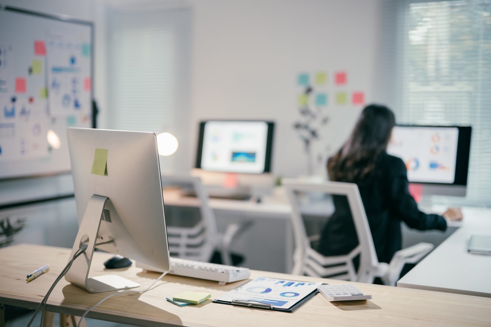 Professional workspace featuring sleek computer setup, office supplies scattered across clean desk surface, female professional typing on laptop in background