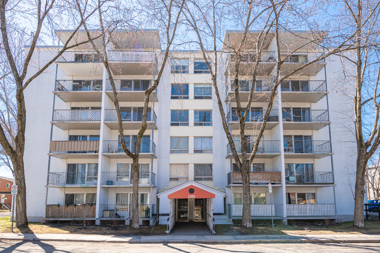 A six-story residential building with balconies, surrounded by leafless trees on a clear day.