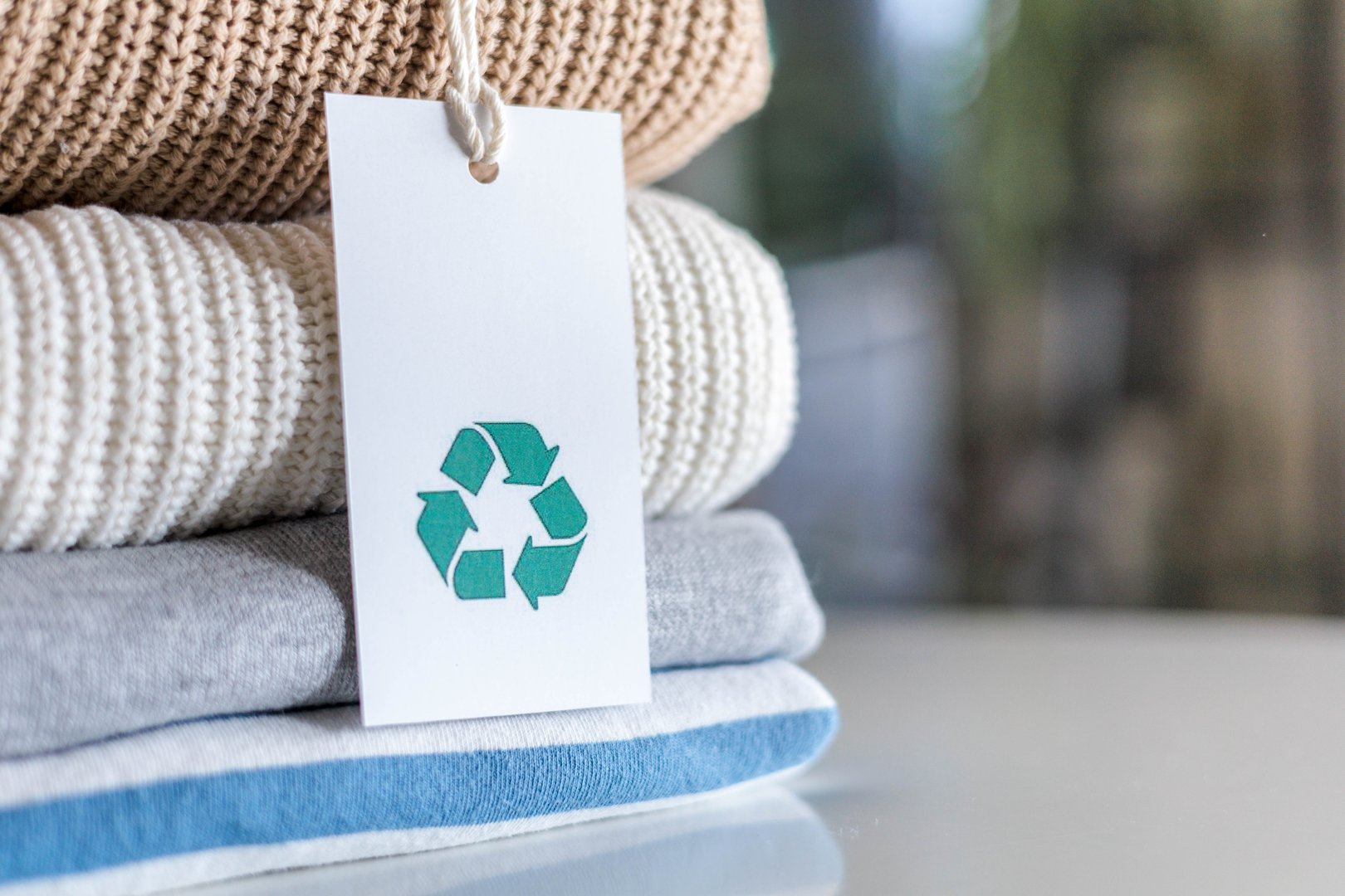 Knitted warm clothes neatly folded heap with craft tag recycling material circle arrow closeup. Eco friendly fashion clothing laundry garment made by recycled zero waste renewable textile technology