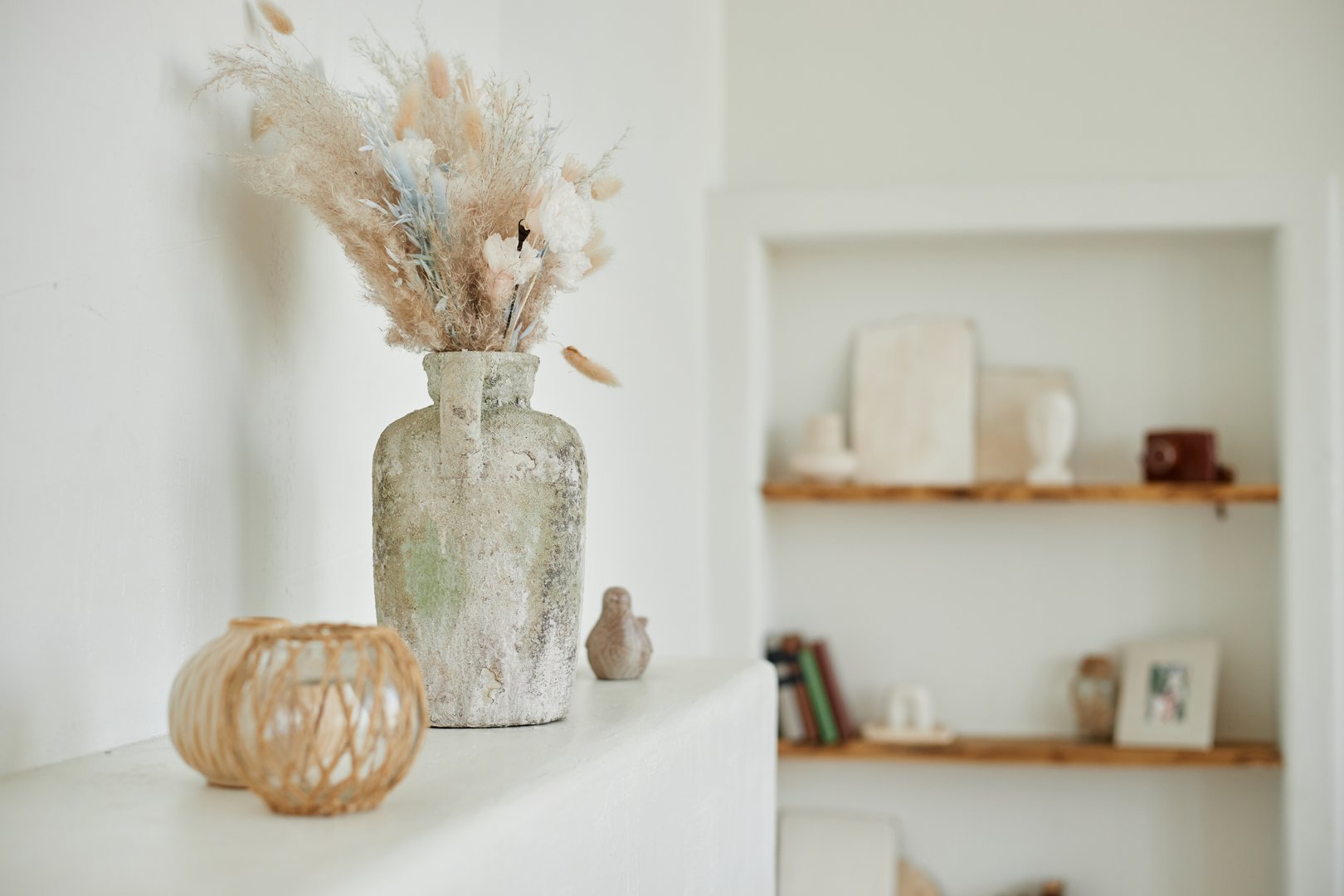 Still life showing rustic ceramic vase with dried grass arrangement standing on ledge, small woven candle holder and decorative bird figurine placed nearby, blurred shelves in background