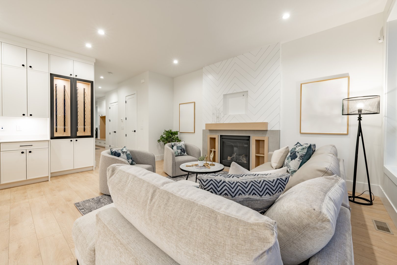 Living room interior of a new construction home in a residential neighborhood, the photos show the blue hour twilight through the windows. The family home features comfortable upholstered furniture a fireplace and simple decor in an open concept space with a view from the living room to the kitchen and dining room. Hardwood floor has an area rug in the middle.