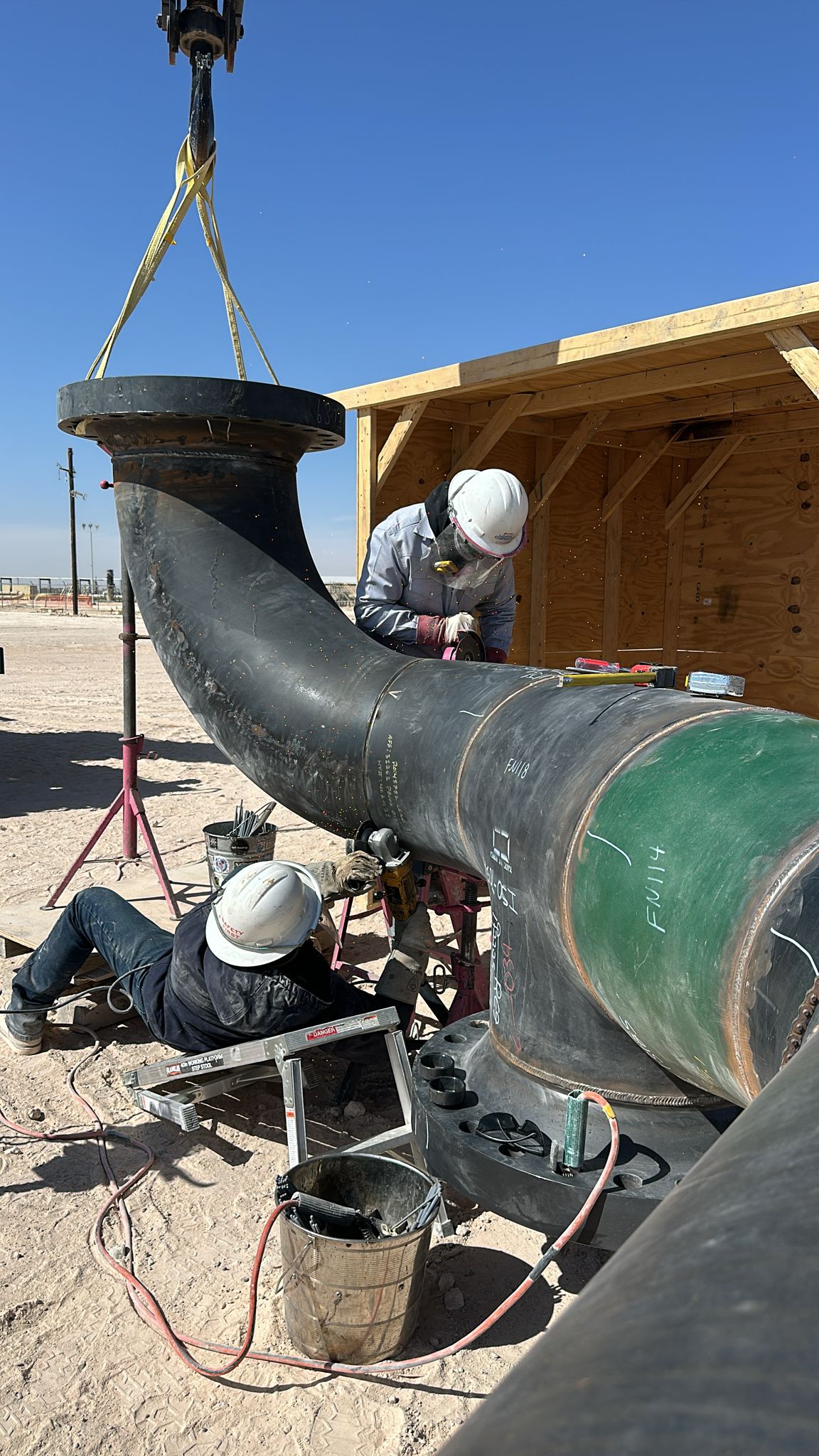 Workers in hard hats weld large pipes at a construction site in a desert setting with a wooden shed in the background.