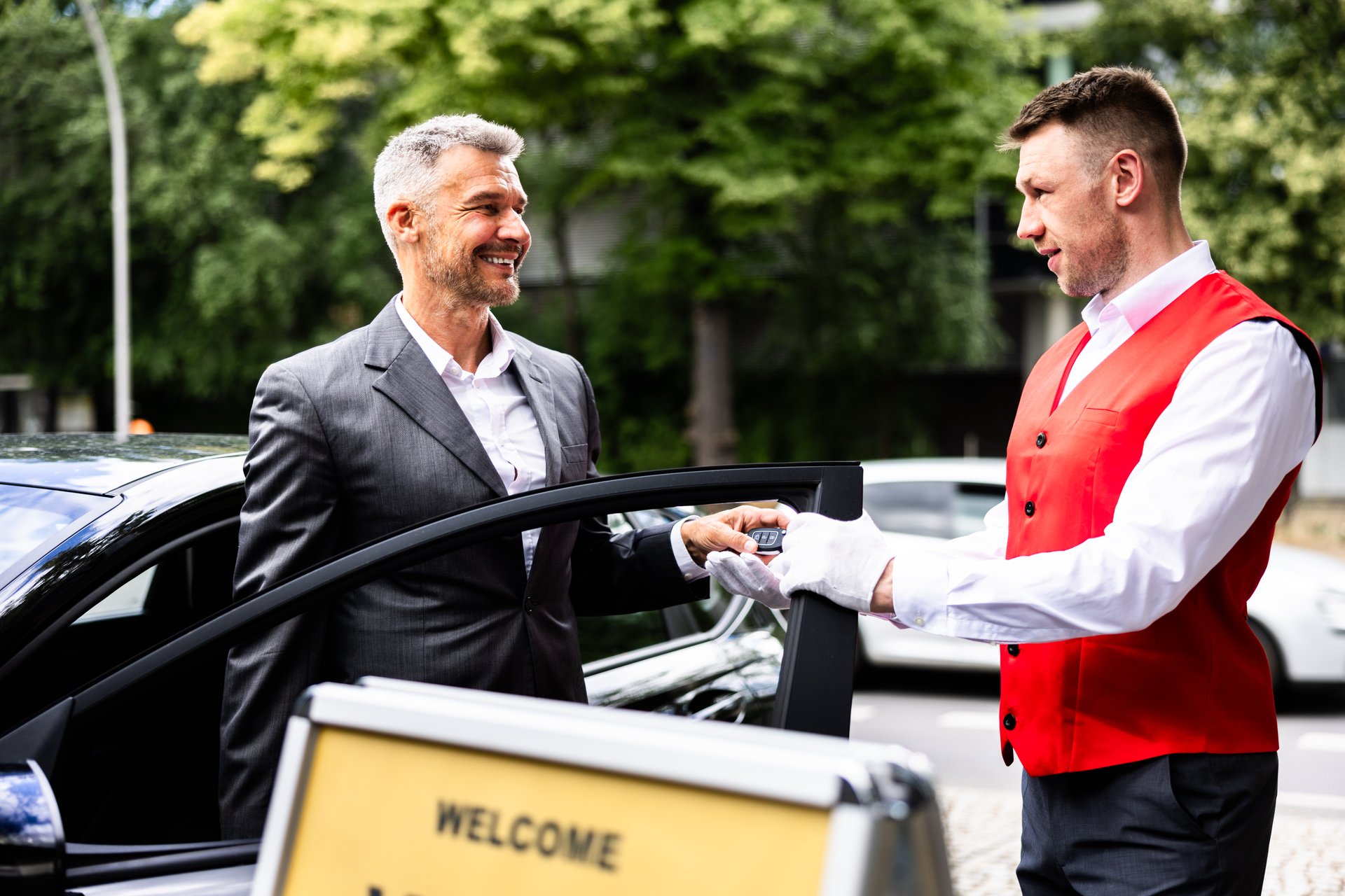 Premium Car Rental Service: Professional Businessman Smiling as Valet Hands Over Keys beside Stylish Vehicle on Sunny City Street.