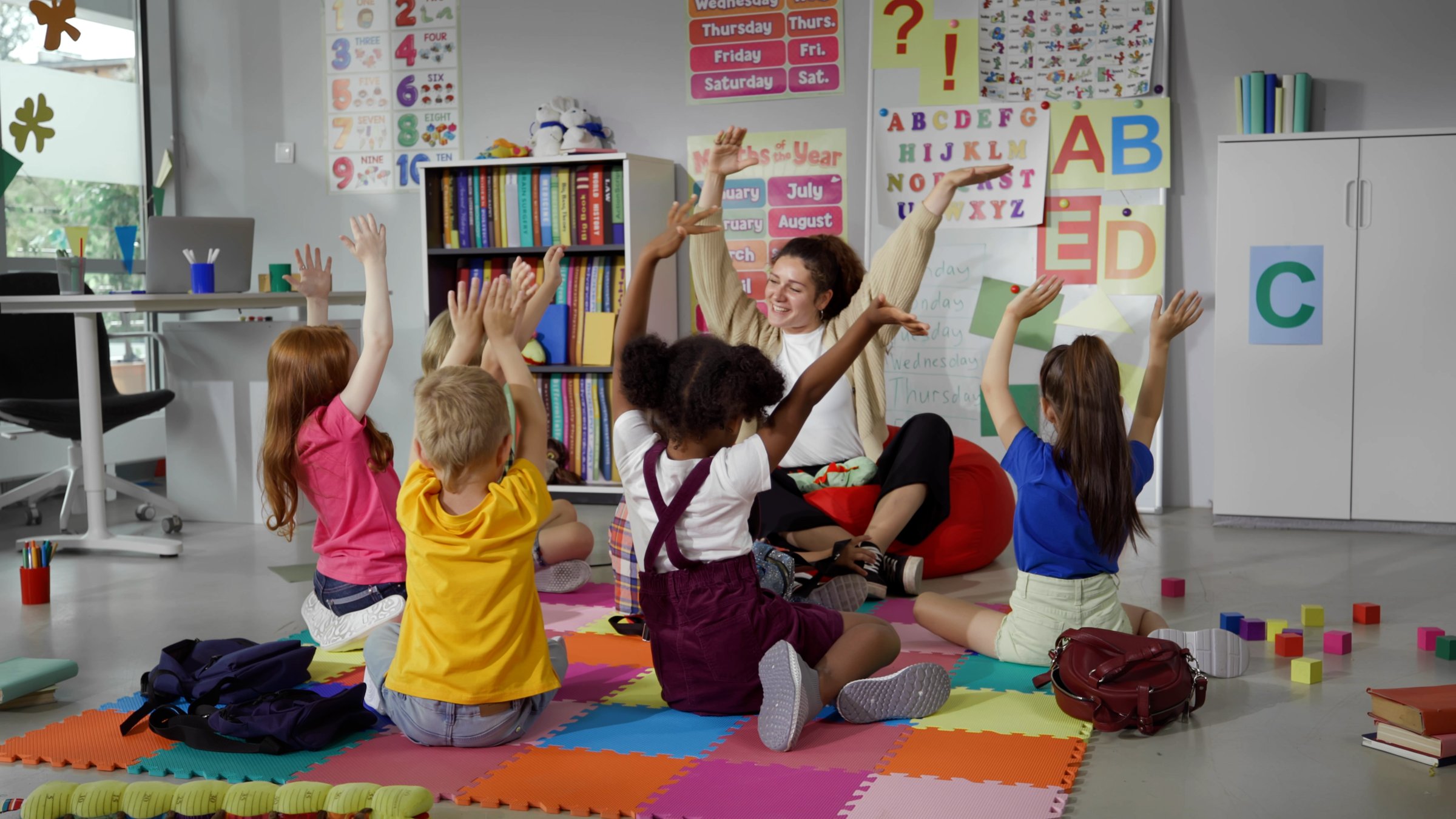 Elementary school teacher sit in class on floor with little pupils and moving hands up. Multiethnic kids with teacher in kindergarten