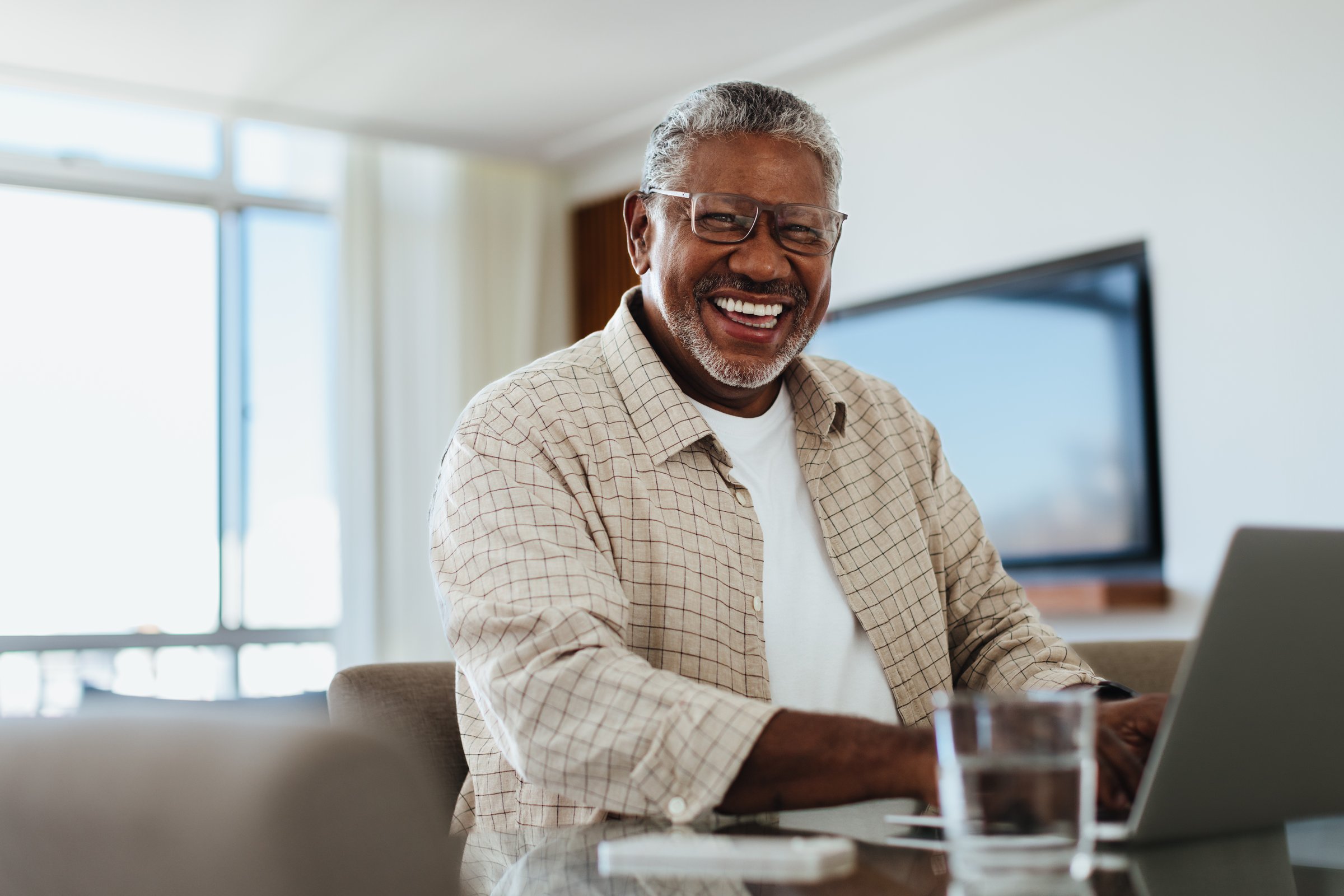 A joyful older male is smiling while using a laptop at home, capturing a happy moment of relaxation and contentment.