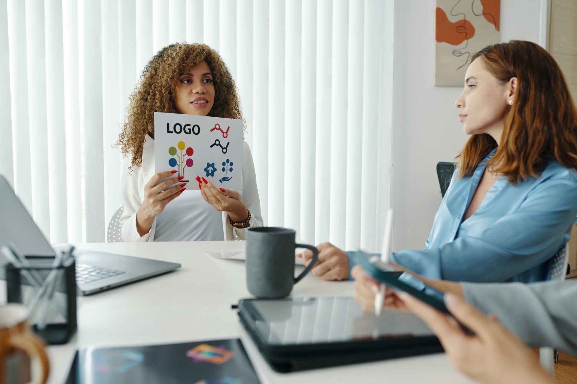 Curly-haired woman showing logo design to seated colleagues in modern office setting with laptops and coffee cups on table