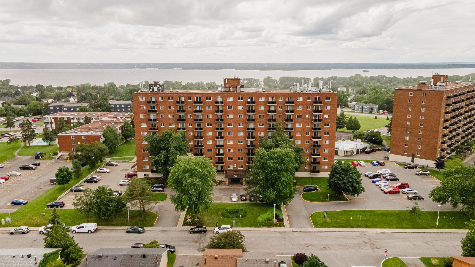 Aerial view of a brick apartment building surrounded by trees, parking lots, and roads; river and greenery in the background.