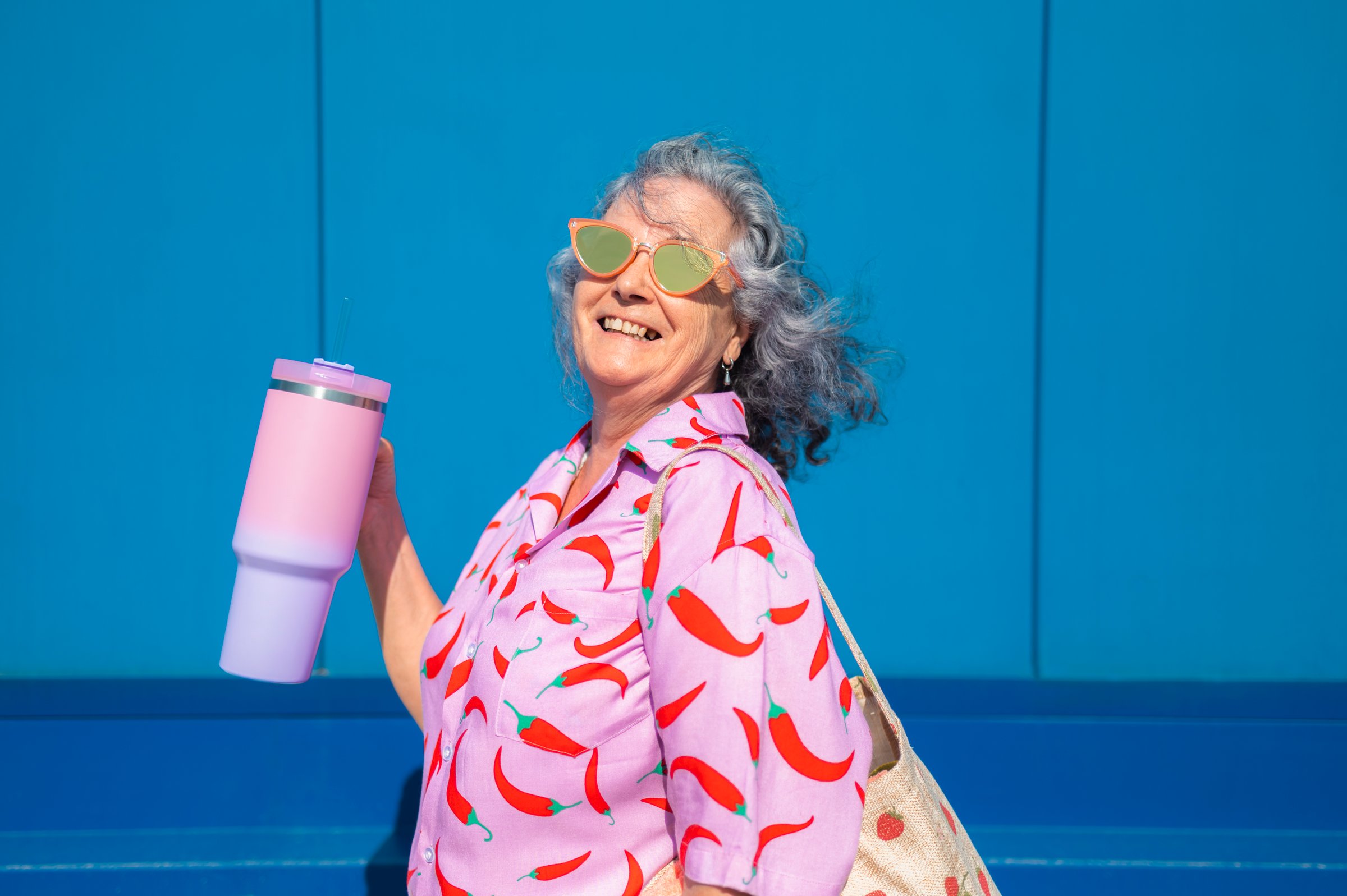Happy senior woman with pink shirt and sunglasses holding a pink tumbler with straw and smiling on a blue background
