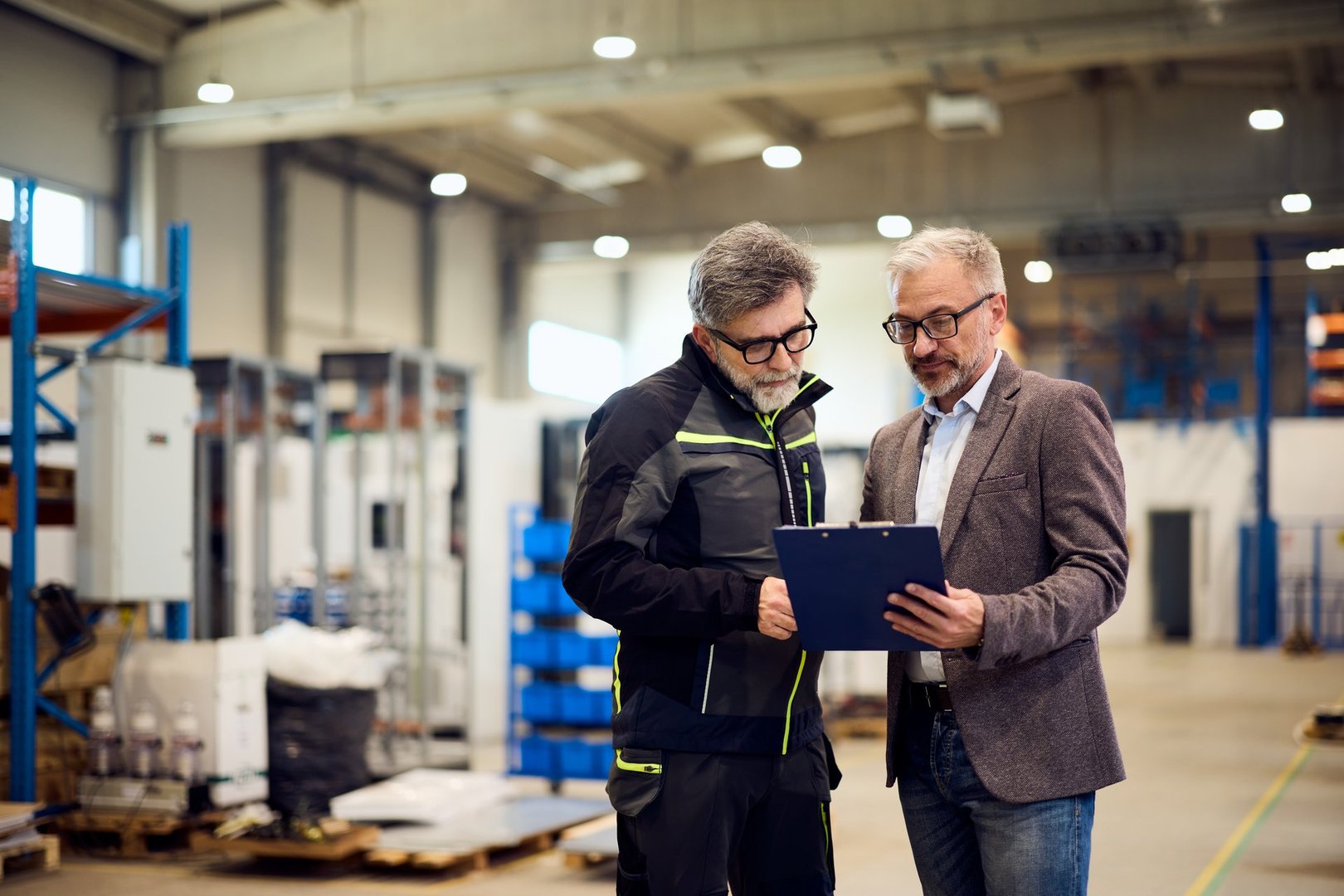 Two mature professionals reviewing documents inside a large industrial or warehouse facility.