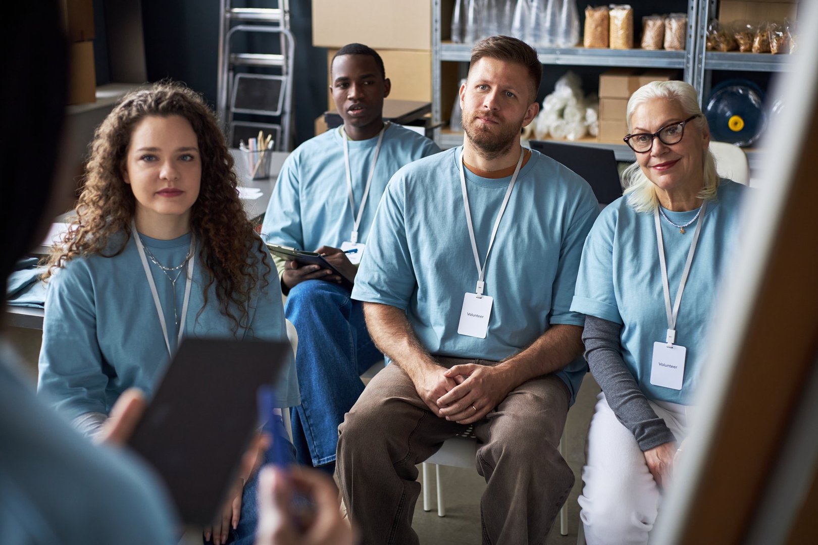 Group of diverse individuals attentively listening to a speaker at workshop. Participants wearing blue shirts and name tags, seated in organized setting with shelves in background