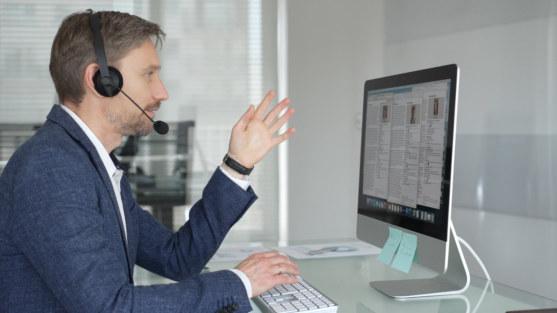 Male professional wearing a headset, communicating virtually on a computer, providing customer service in a modern office setup. Business people concept