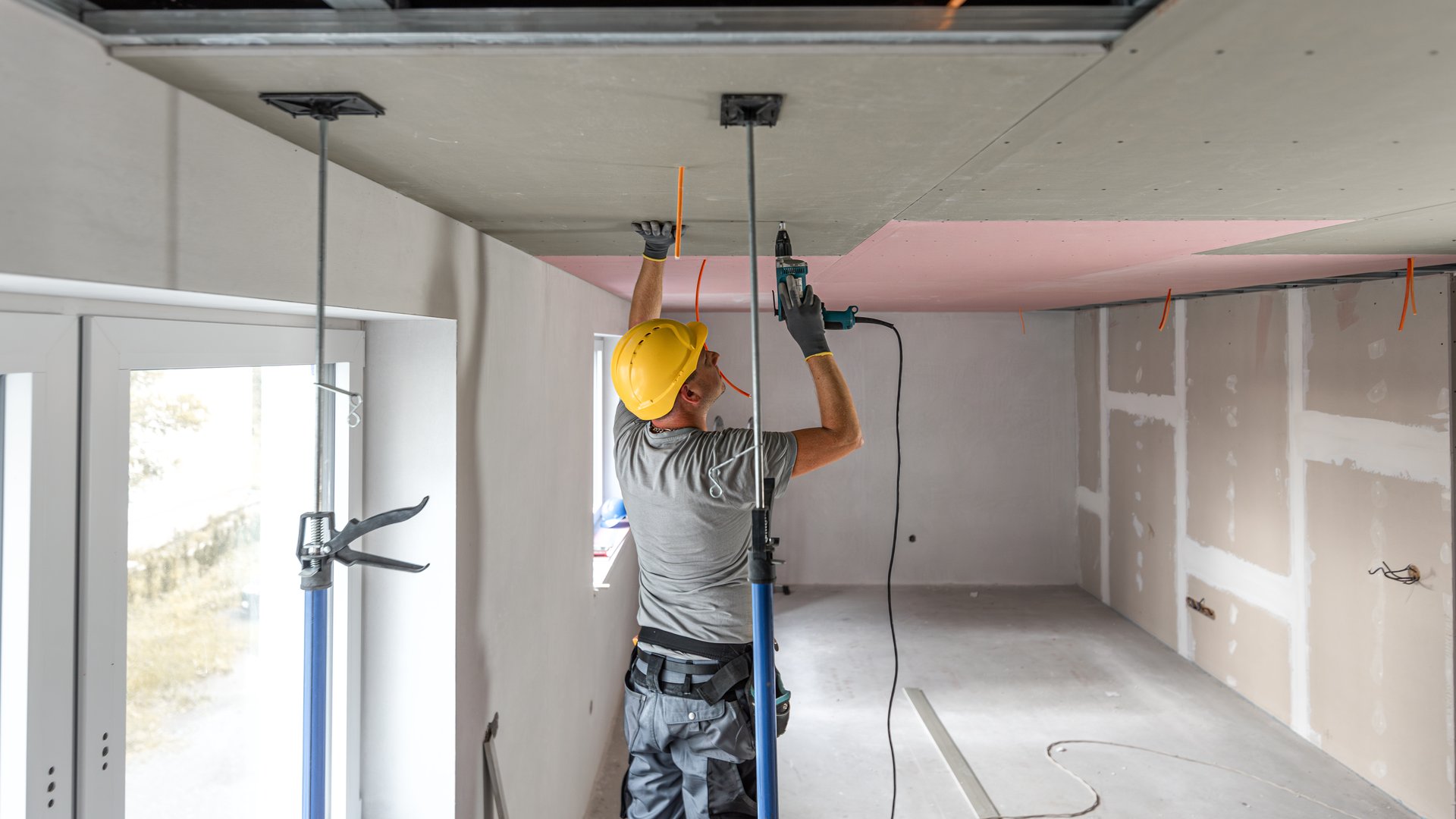 The worker screwing plasterboard to the ceiling. He is using special electric screwdriver and drywall lift. A red plasterboard improve the fire resistance of ceiling structures.