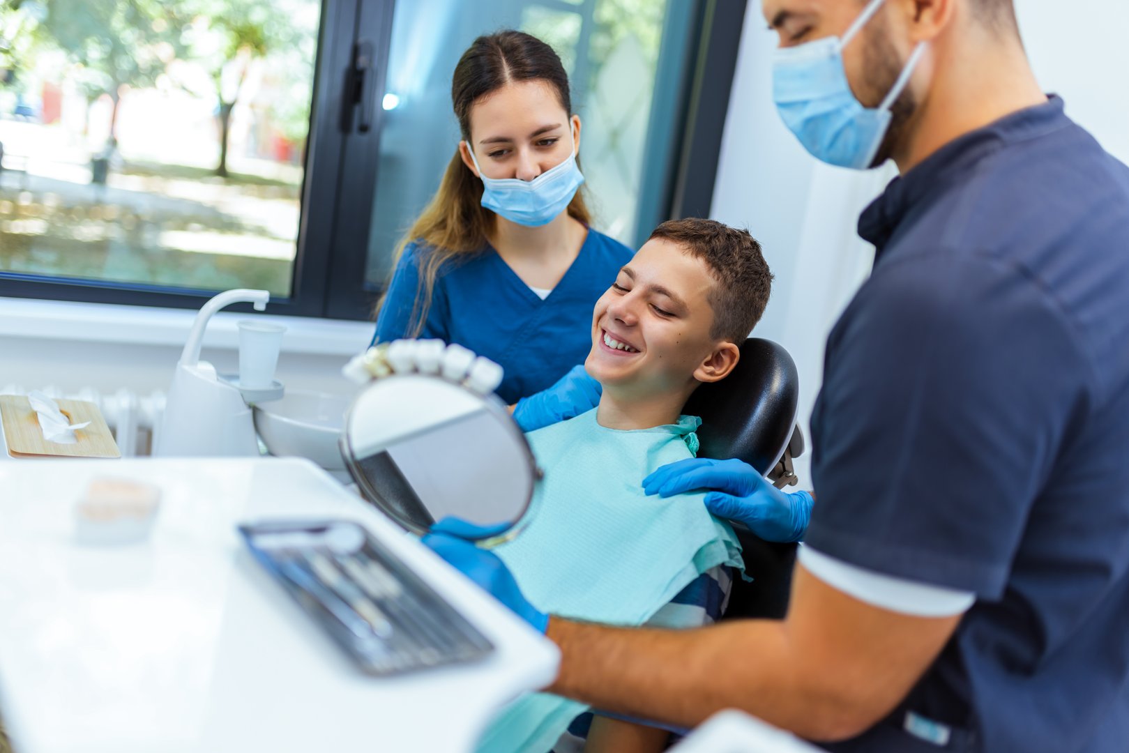 Little boy sits in dental chair, smiling happily as doctor and nurse hold a mirror to show his teeth, demonstrating joyful pediatric dental care in a modern clinic.