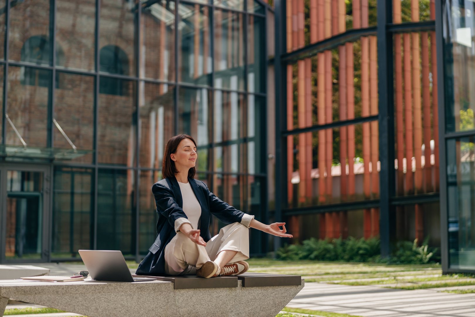 Relaxed businesswoman meditating in outdoor office area, embracing work-life balance, laptop nearby.