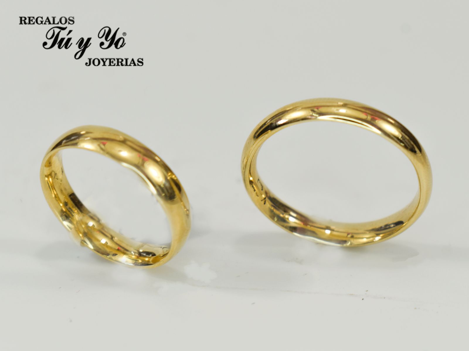 Two gold rings with a polished finish on a light background, featuring "Regalos Tú y Yo Joyerías" text in the corner.
