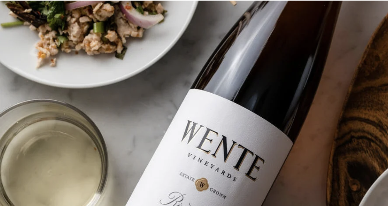 Wente Vineyards wine bottle on a table with a glass of wine and a plate of food.
