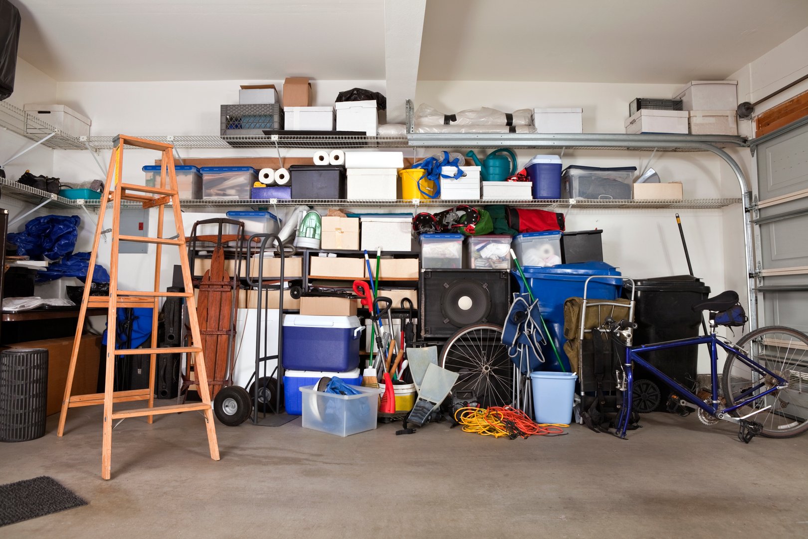 A cluttered garage with shelves stacked with boxes and storage bins, a wooden ladder, bike, and various tools and items.