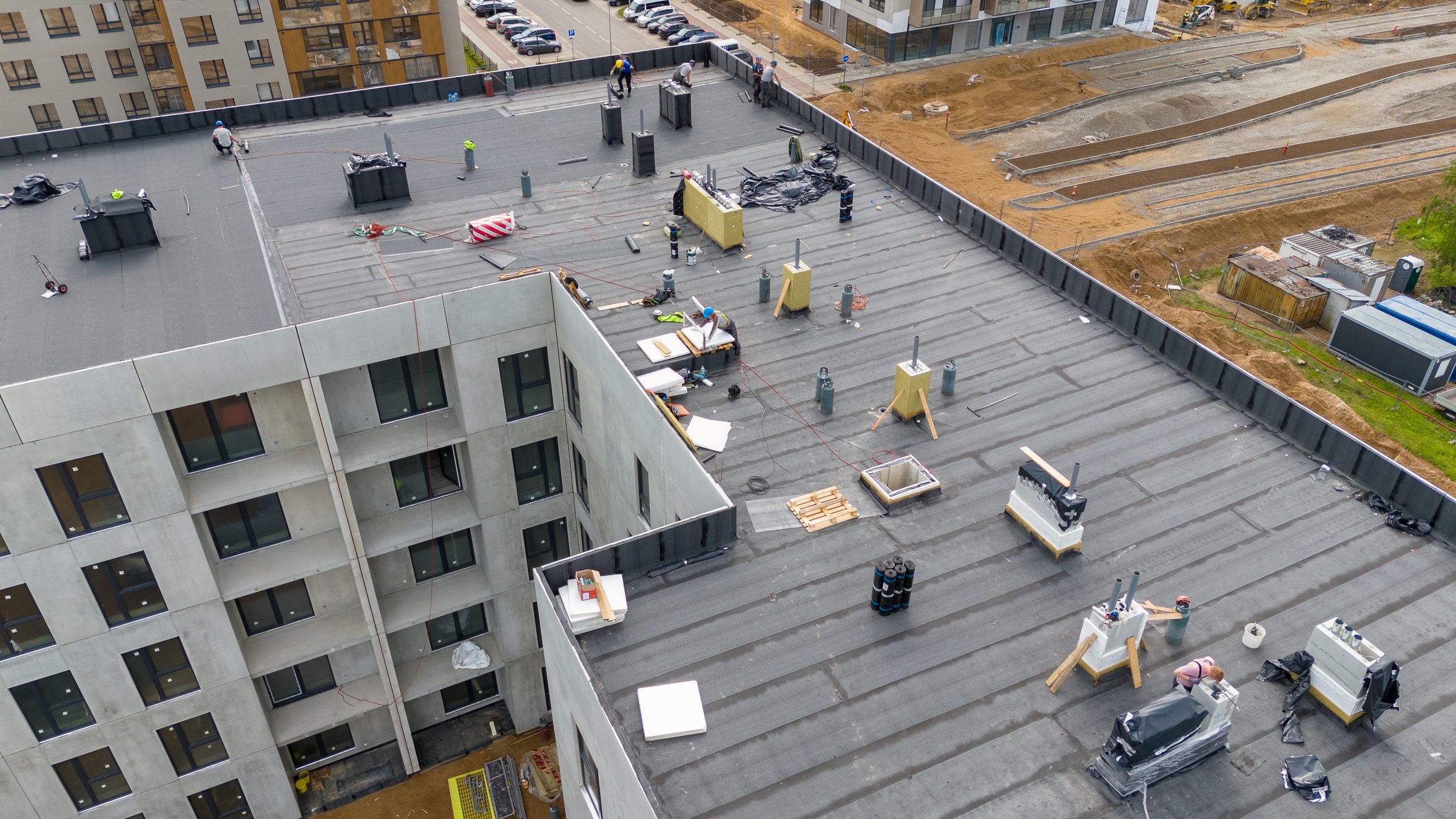An aerial view of a construction site featuring workers on a flat roof. Various construction materials and equipment are scattered across the roof, with nearby buildings and a construction area.
