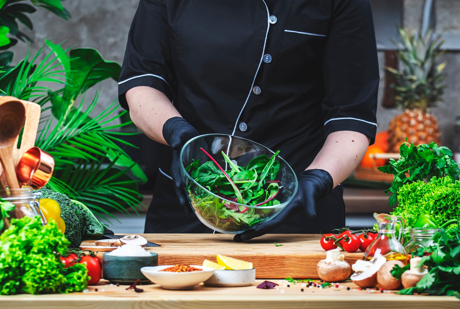 Chef holds bowl of salad with arugula and chard in hands. Cozy kitchen with wooden table, kitchenware, vegetables, herbs and ingredients for cooking. Healthy vegan food, eating, culinary, recipes, blogging
