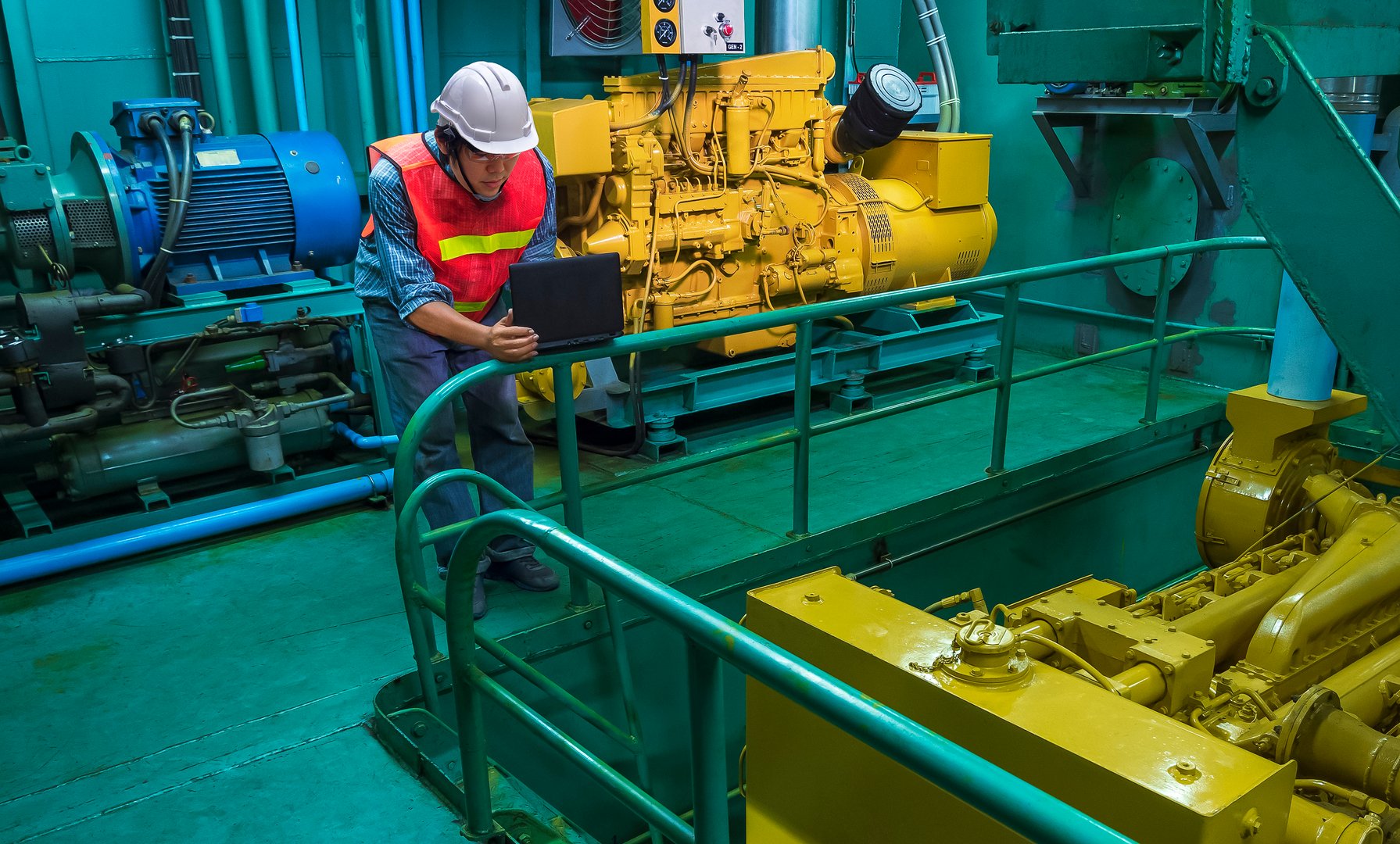 Asian young engineer in reflective clothing with helmet using laptop computer to checking engine quality system inside of engine room of fishing vessel