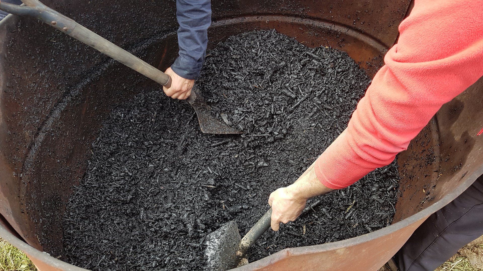 two men with shovel removing charcoal in a drum. use of biochar in the vegetable garden and preparing it.