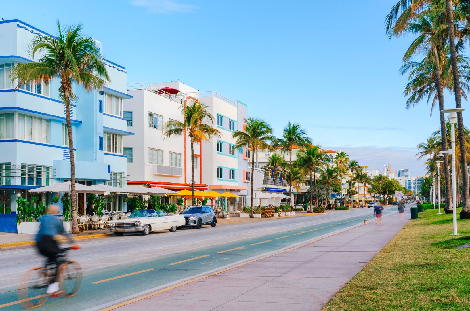 Ocean Drive in Miami Beach, lined with pastel-colored Art Deco buildings, palm trees, outdoor cafés, and a bicycle lane, with people enjoying the morning activities  under a clear blue sky.