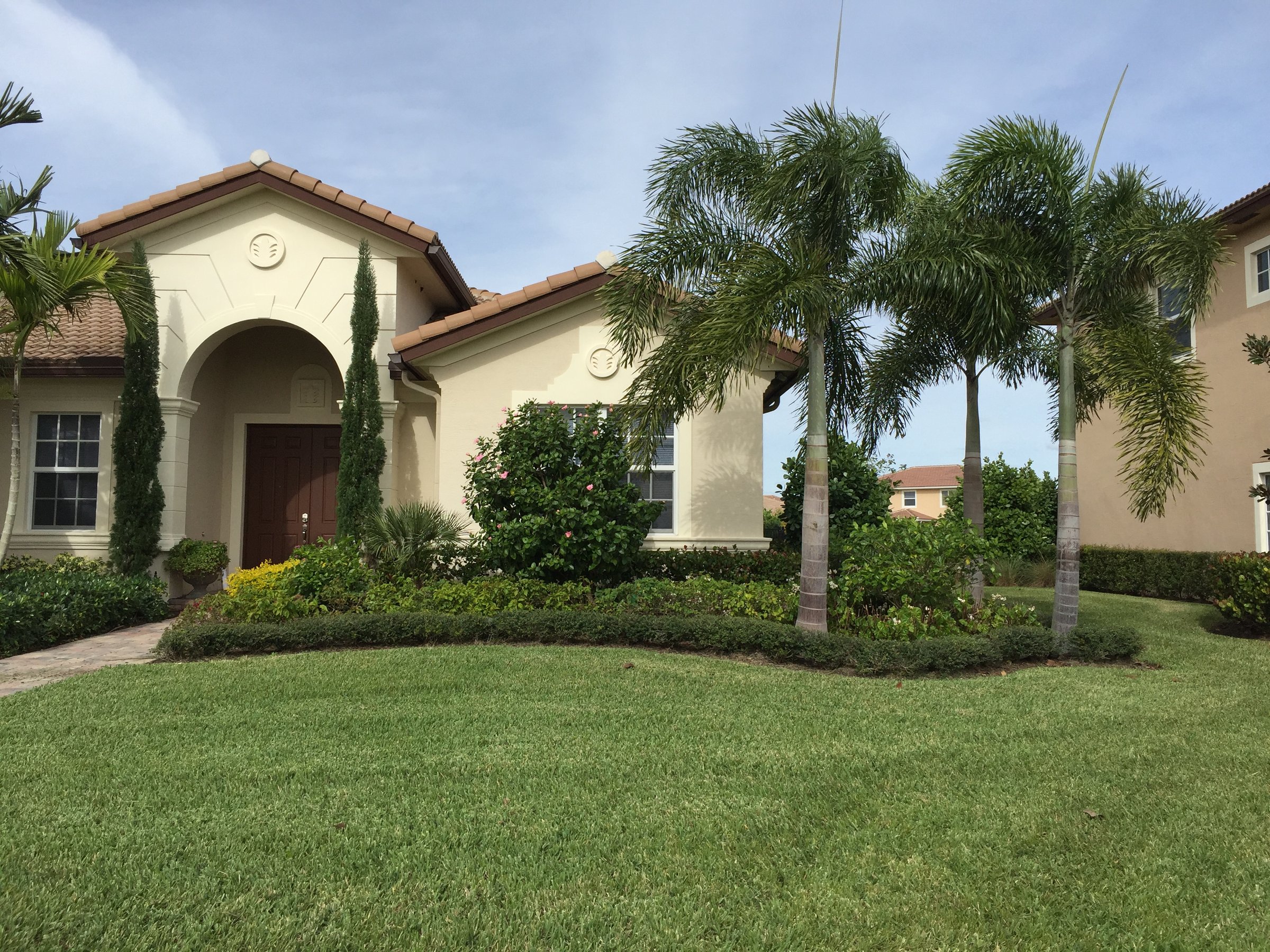 A beige stucco house with a tiled roof, lush landscaping, palm trees, and a manicured lawn under a clear blue sky.