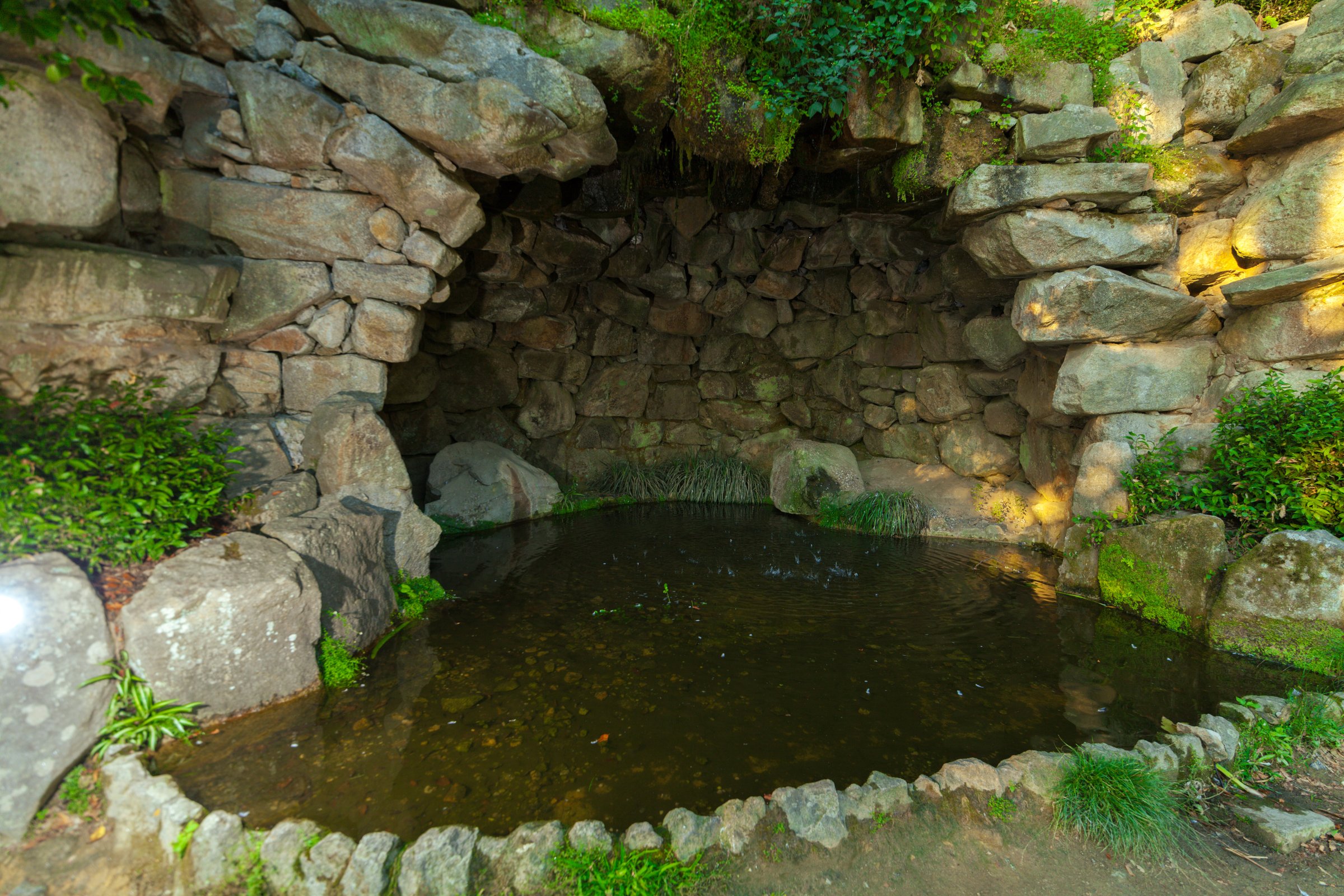 A shaded stone grotto with a small, calm pond surrounded by natural rock formations