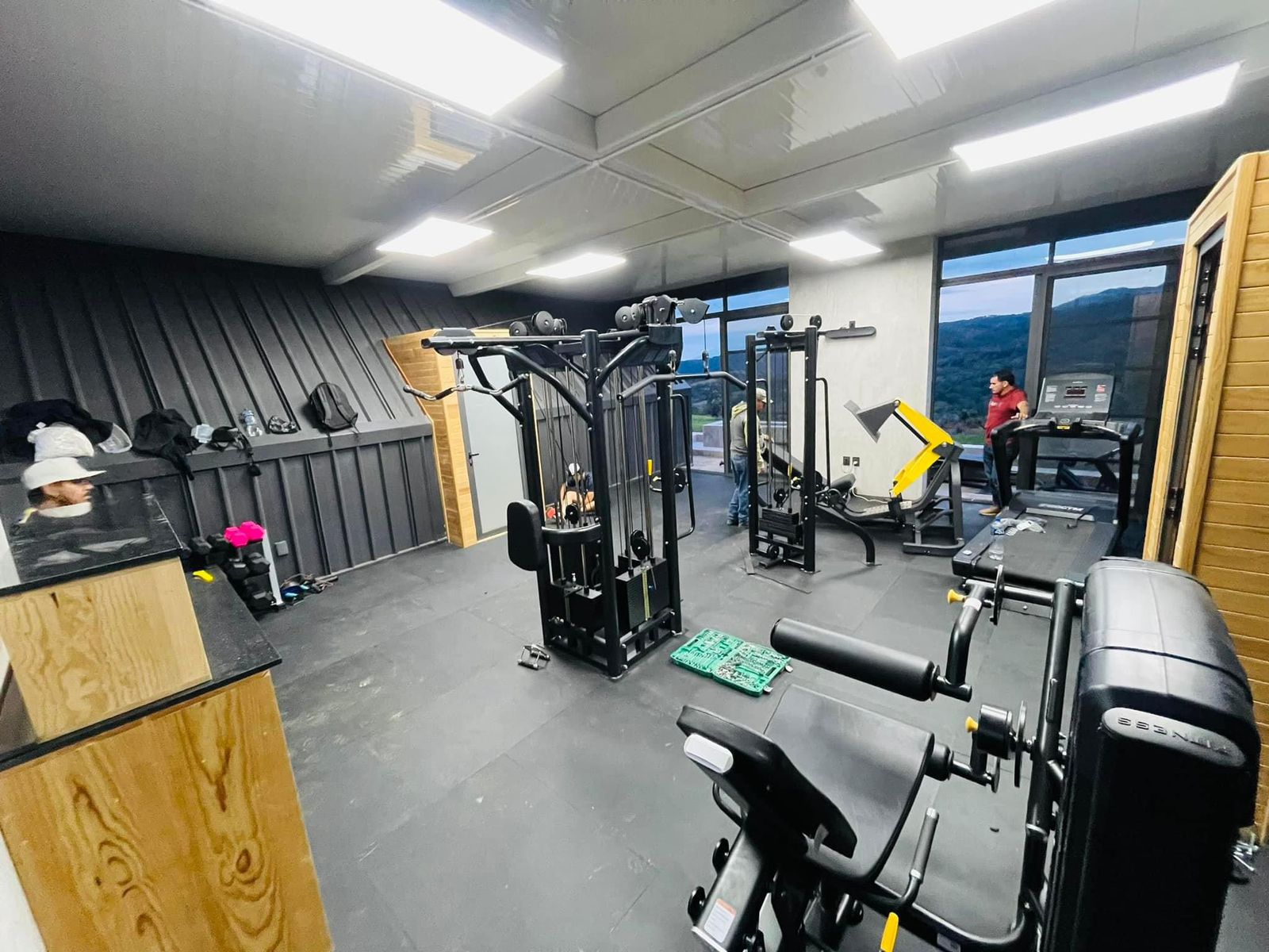A gym with various exercise machines, rubber flooring, and large windows. Two people are present, one working out on a treadmill.