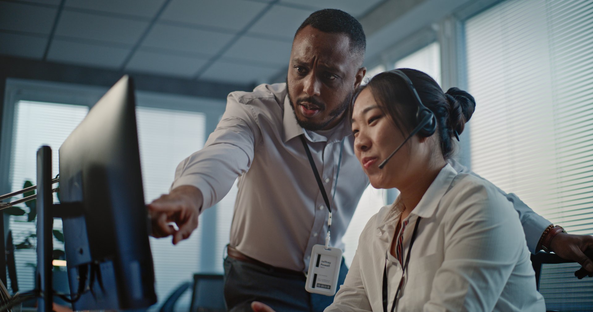 Modern call center office: Asian helpdesk specialist in headset uses computer, talks to African American colleague. Two diverse technical support operators working in online customer support service.