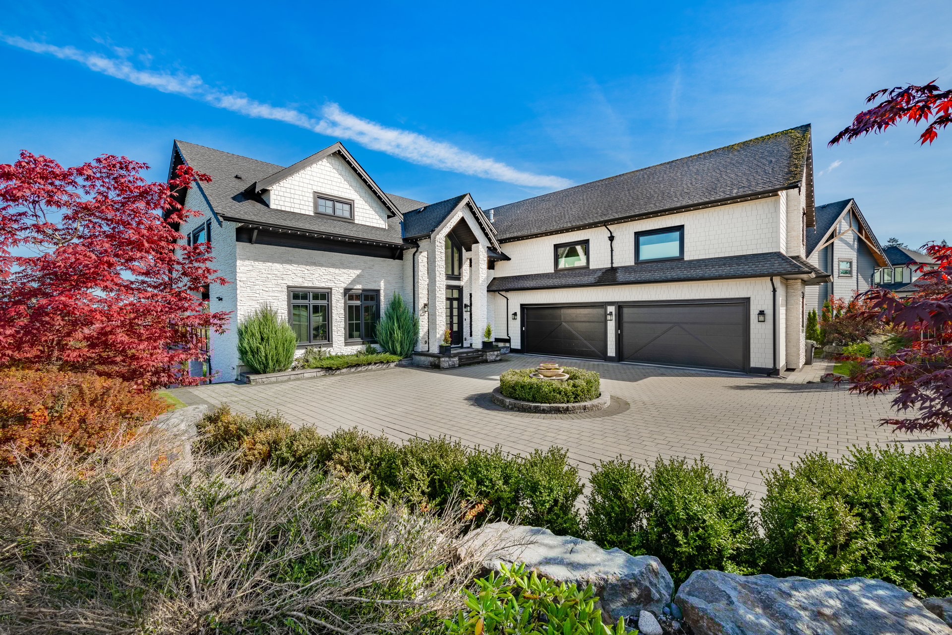Elegant and stately home on a large lot on a sunny autumn fall day blue sky lush green grass colorful leaves on the trees the home is set against a deep blue sky and it is built with light colored stone and black wood trim black roof two large garages brick pillars and a wooden fence around a cobblestone driveway with a fountain in the centre and fully landscaped