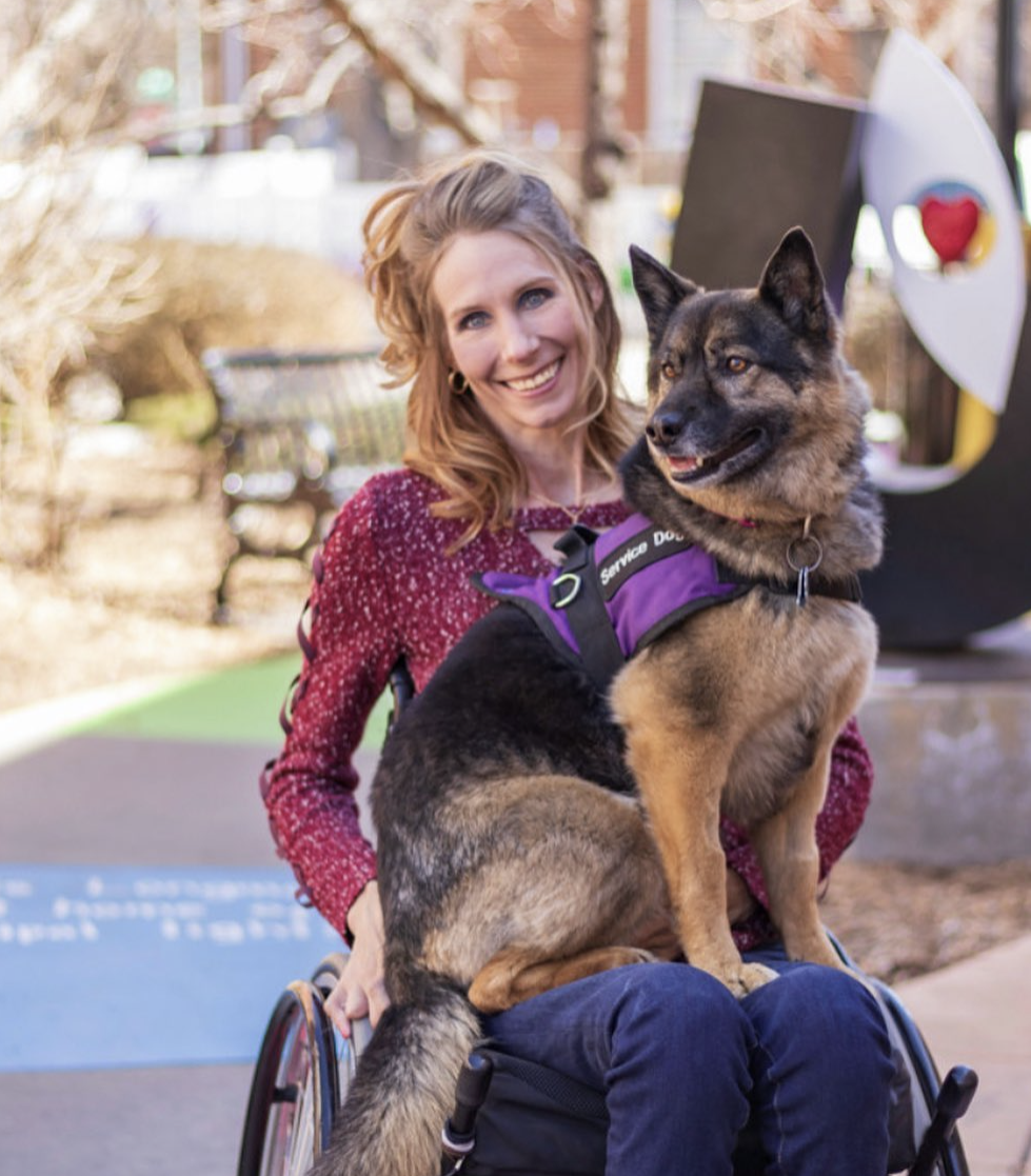 A woman sits in a wheelchair, smiling with a German Shepherd service dog on her lap, outdoors with a statue in the background.