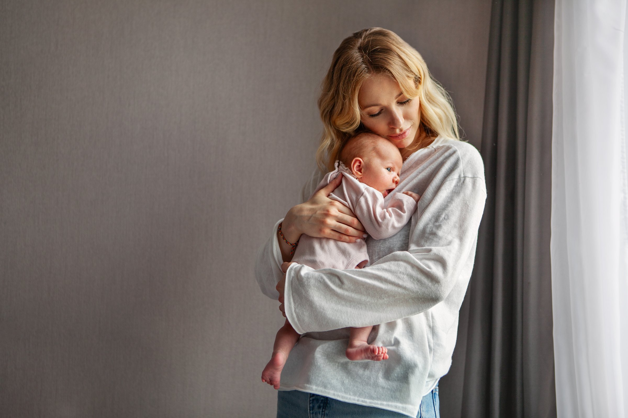 Young beautiful mother holds newborn baby while standing at home near window. tender moment with baby dressed in soft pink And mom in light clothes. Natural light, peaceful and intimate atmosphere