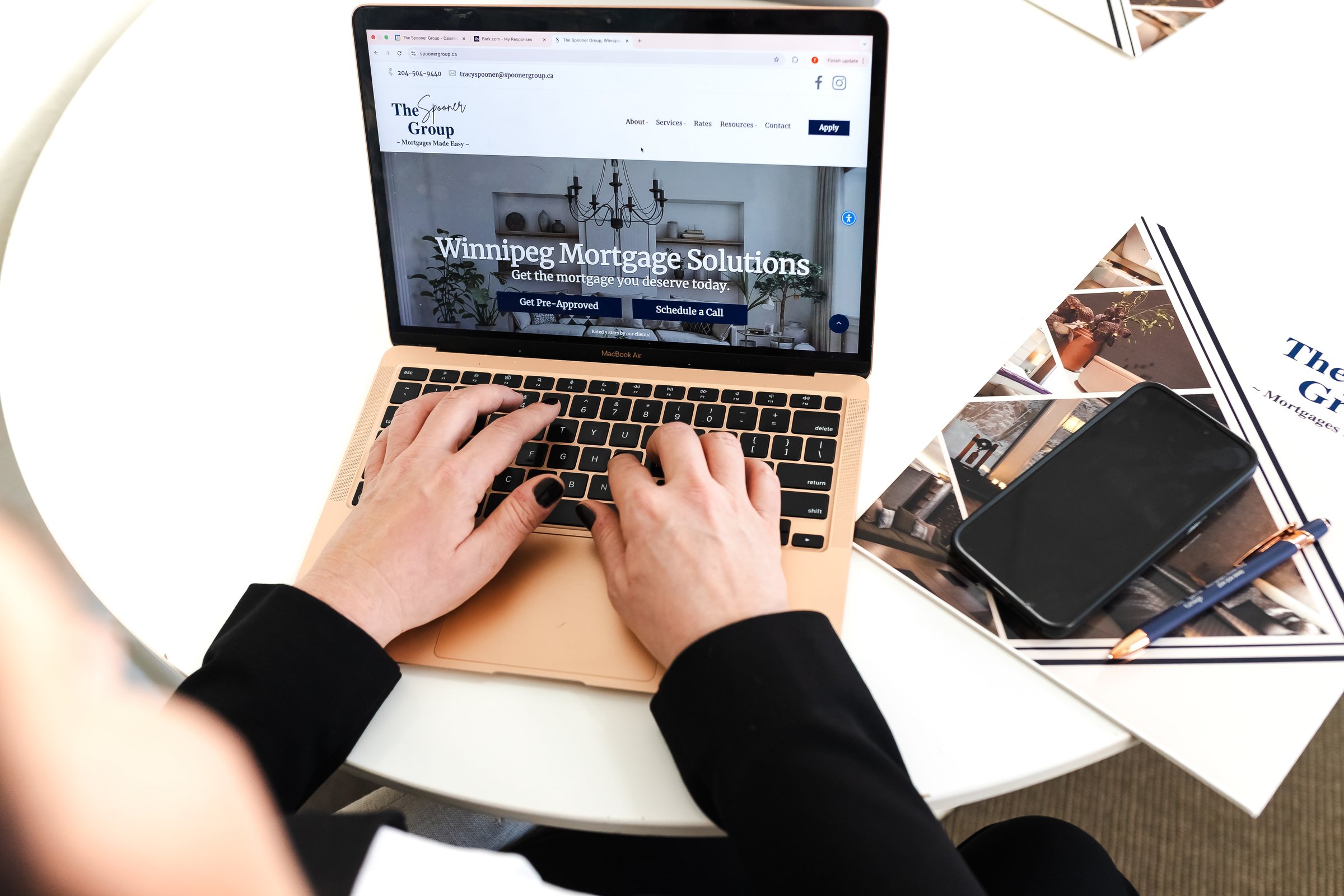 Person using a laptop with "Winnipeg Mortgage Solutions" on the screen, next to a smartphone and brochures on a table.