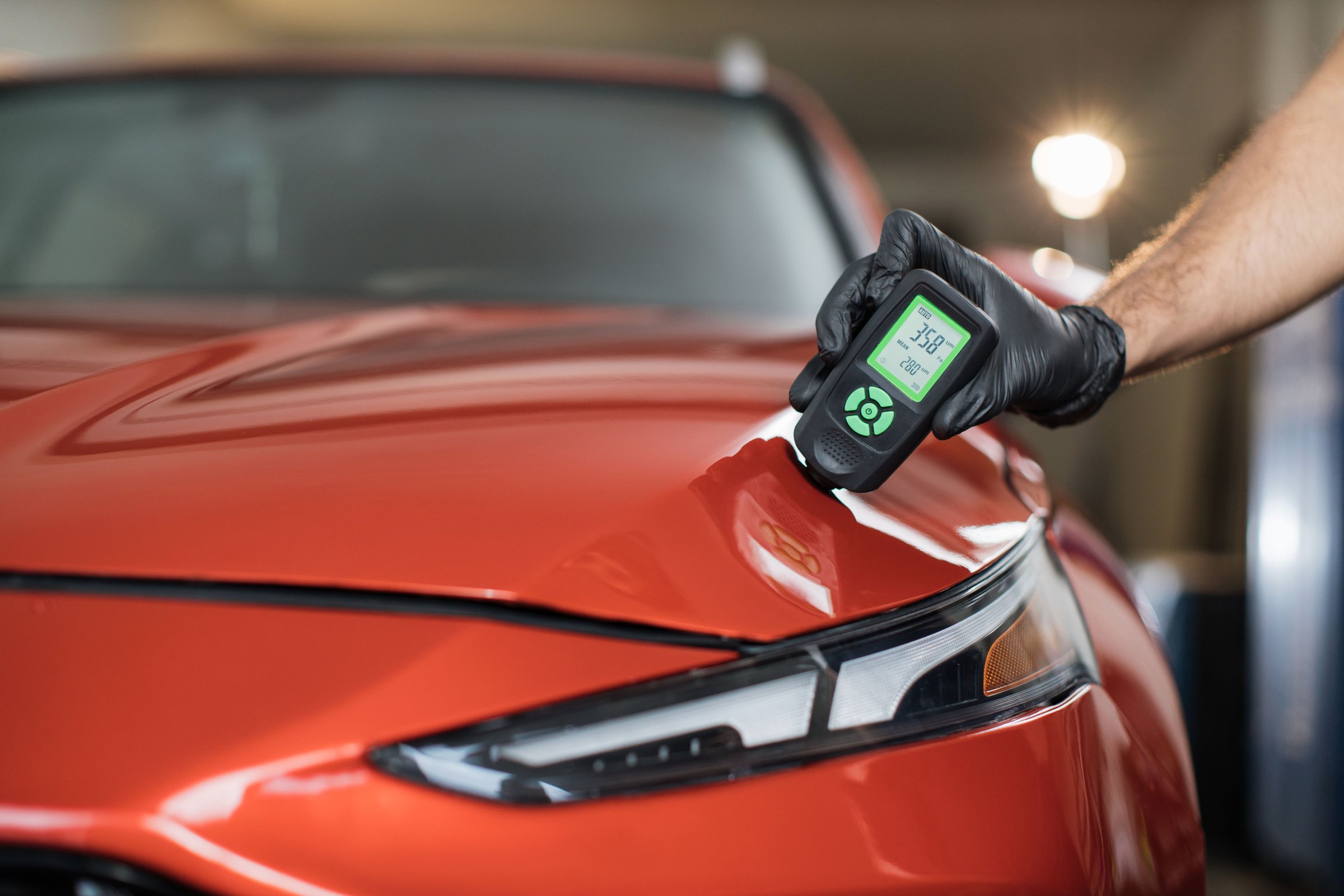 Inspector measuring paint cover thickness of red car body at the vehicle detailing service, close-up view on hand and professional thickness meter