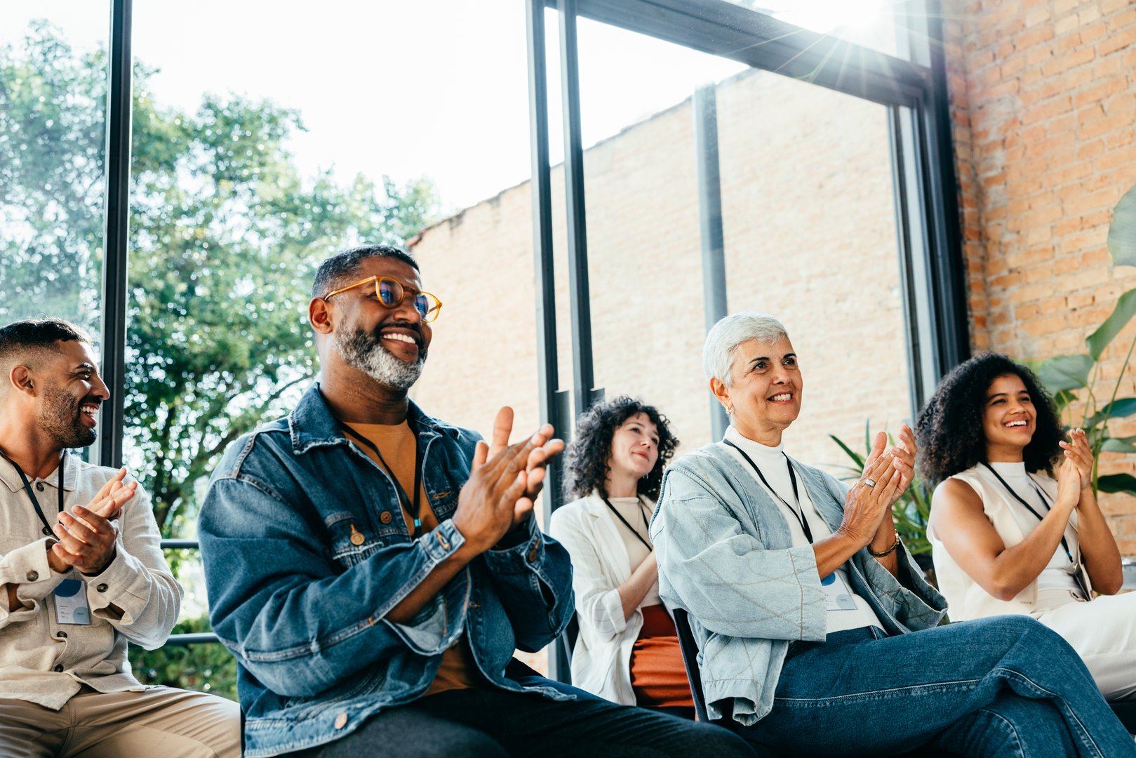 Diverse group of people attending a business meeting, happily clapping and smiling. They are seated in a bright room with large windows and natural light.