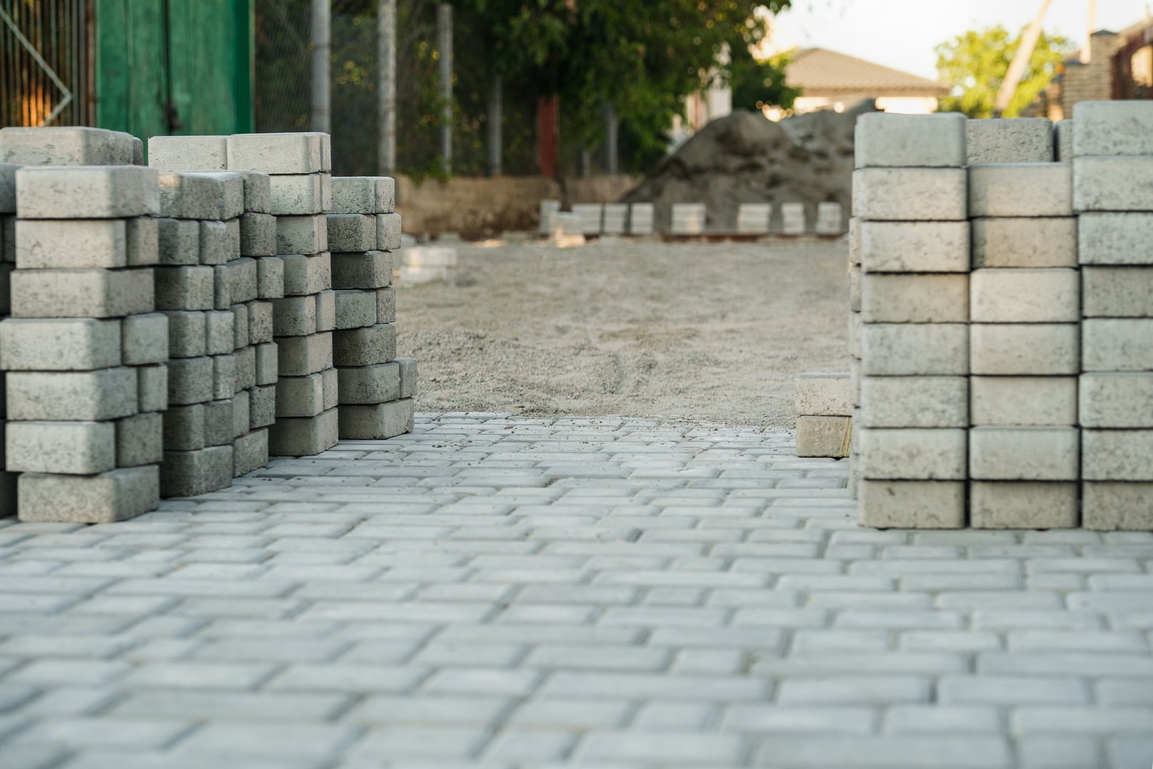 The ground showcases a neatly arranged pathway of gray paving stones, seamlessly leading towards a construction site. Lush greenery and a clear blue sky enhance the setting.