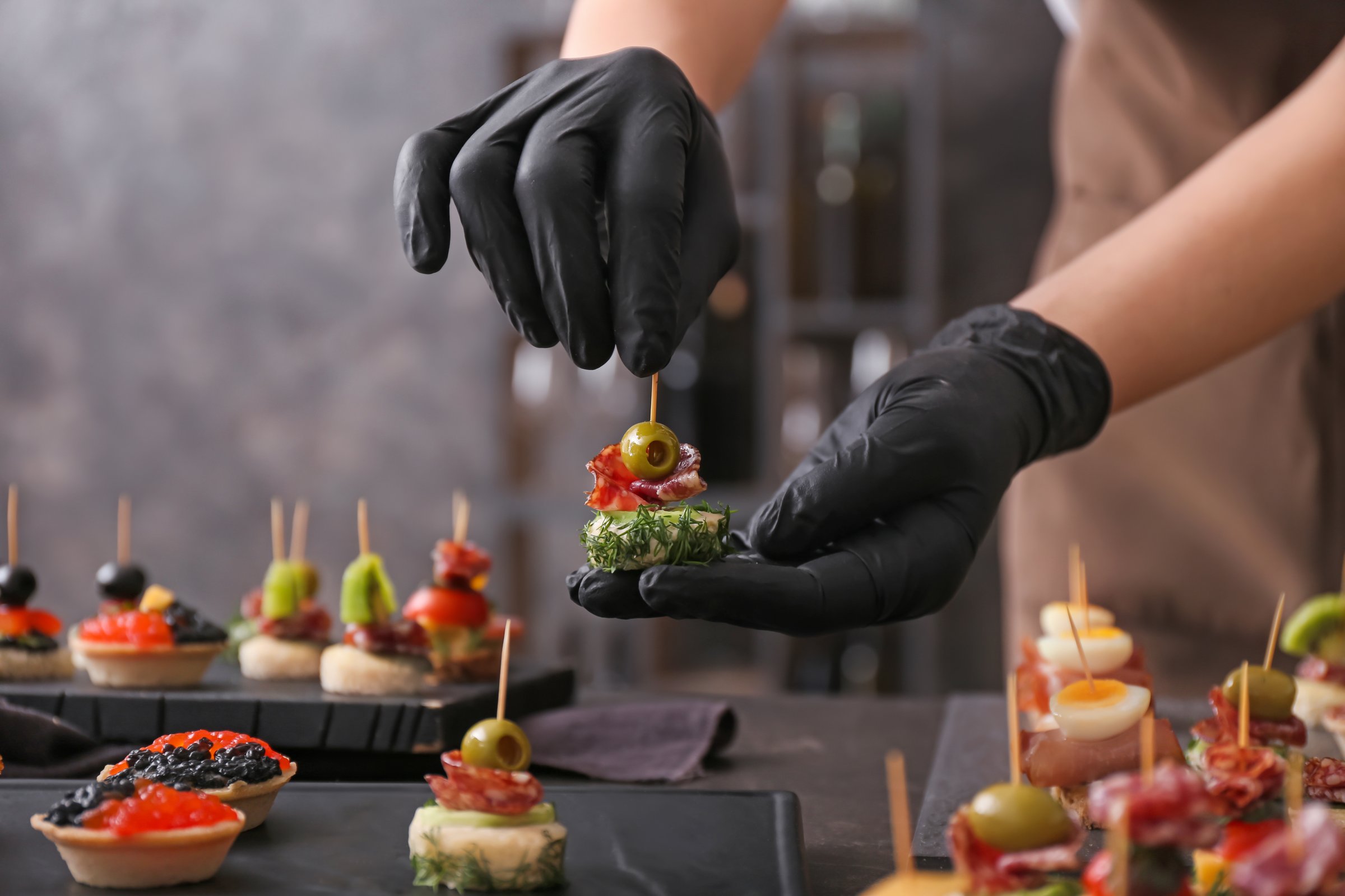Close-up shot showing the hands of a professional chef or caterer, wearing sleek black gloves, precisely placing a garnish on a miniature appetizer stick. Various other beautifully prepared tartlets and snacks are visible in the background, ready for service. This image conveys high-quality catering, professional service, event planning, and fine dining preparation for corporate or private parties.