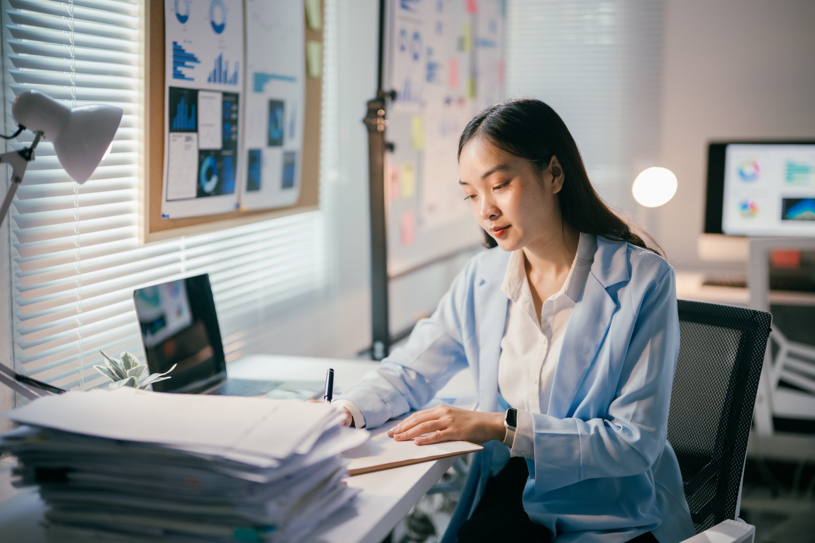 Young Asian businesswoman works late in her brightly lit office, diligently taking notes from a substantial stack of documents on her desk