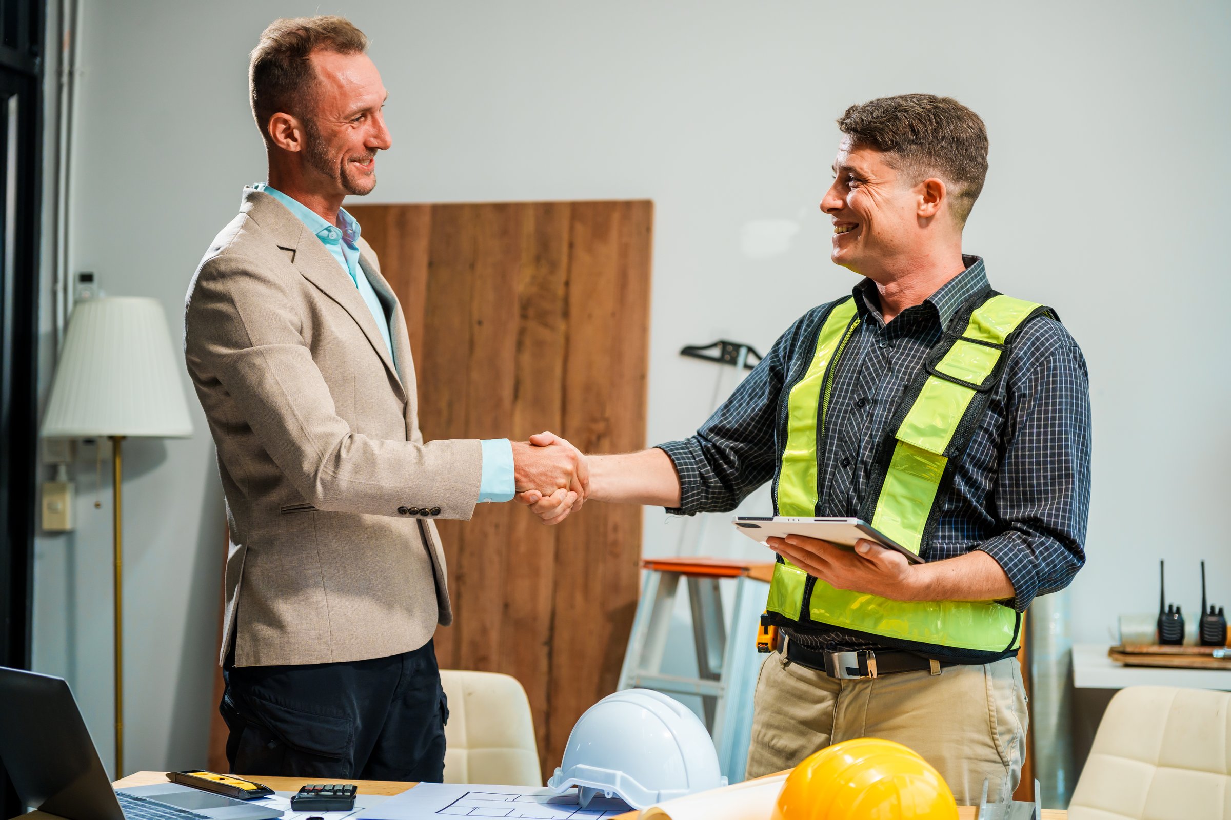 A Caucasian male engineer and an Italian male building contractor are planning and talking at a desk, examining house plans and a scale model with wooden textures for a renovation project.