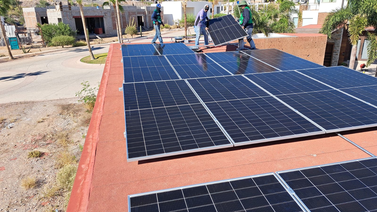 Workers installing solar panels on a red roof in a residential area with trees and unfinished buildings nearby.