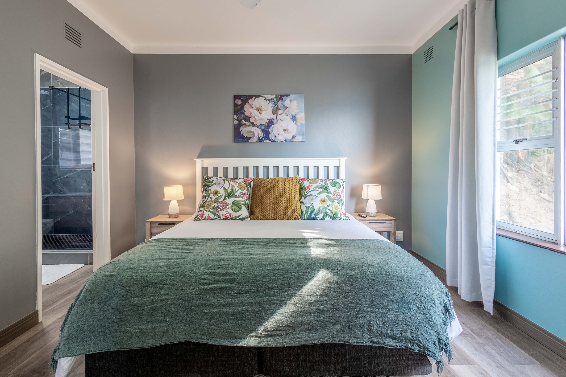 Bedroom with a large bed, colorful pillows, green blanket, bedside lamps, floral wall art, and a window illuminating the space.