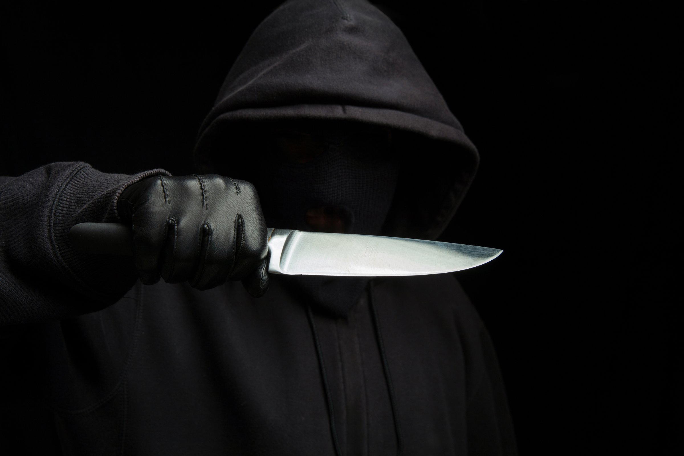 Criminal holding a knife, hooded man holding a weapon in gloved hand, black background, armed attack concept.