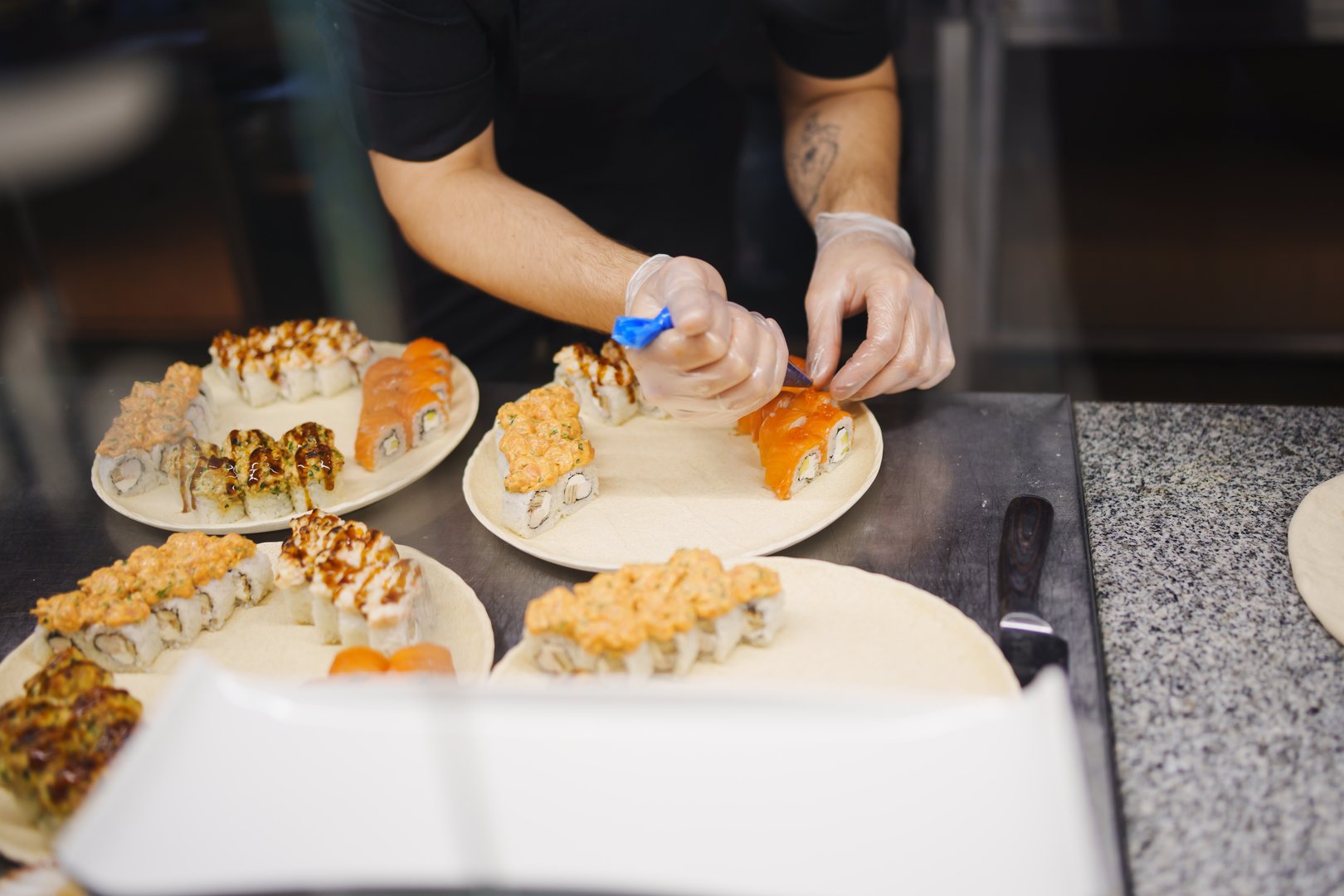 In a bustling kitchen, a chef carefully decorates plates of sushi rolls with vibrant toppings. The focus is on the precision and skill involved in sushi preparation.