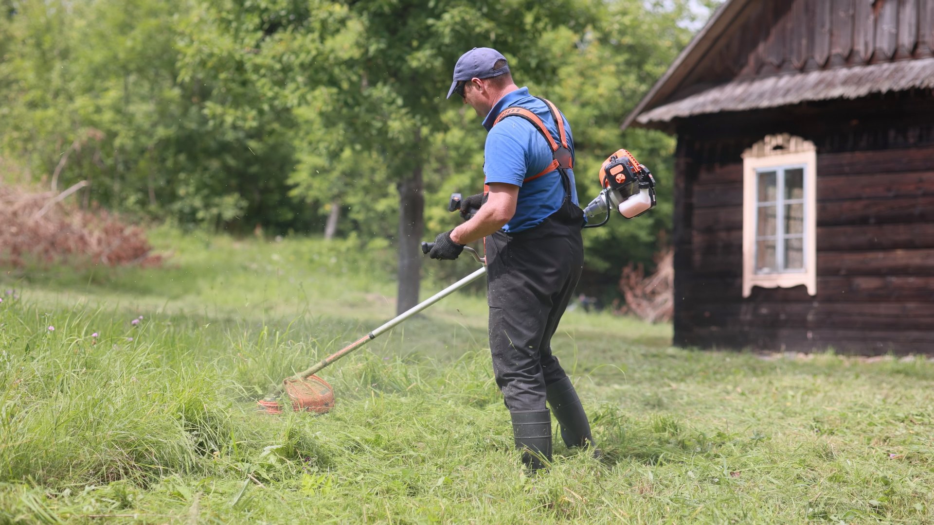 Gardener using brushcutter mowing grass near wooden house in countryside