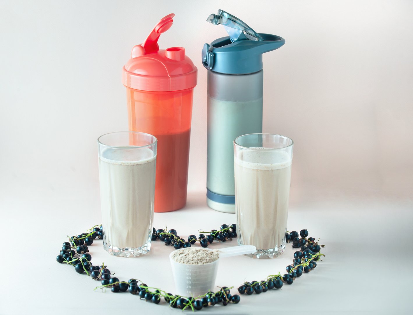 On a light background, two glass glasses with a ready-made protein drink, a black currant heart is laid out around them, shakers in the background