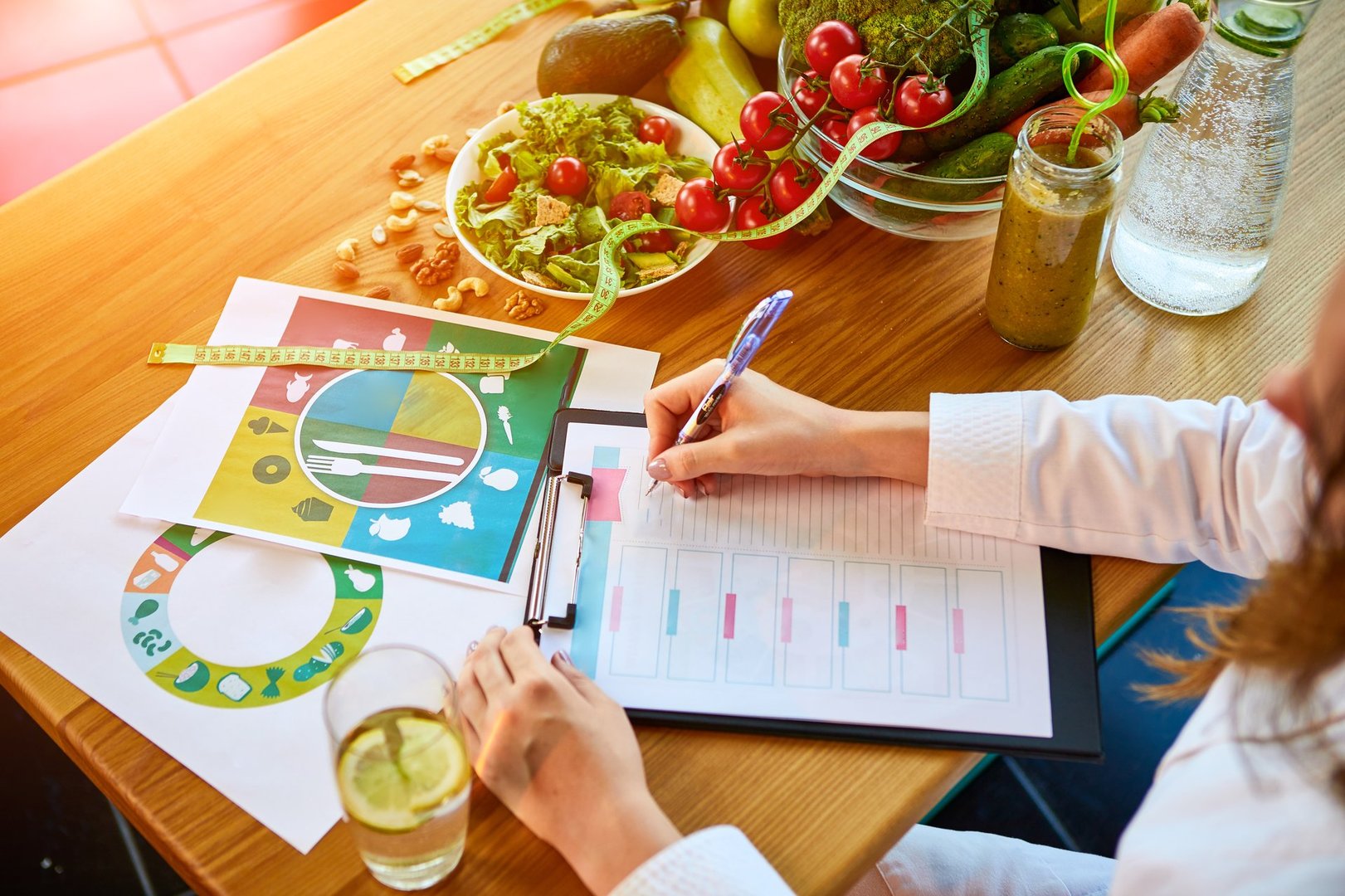 Woman dietitian in medical uniform with tape measure working on a diet plan sitting with different healthy food ingredients in the green office on background. Weight loss and right nutrition concept