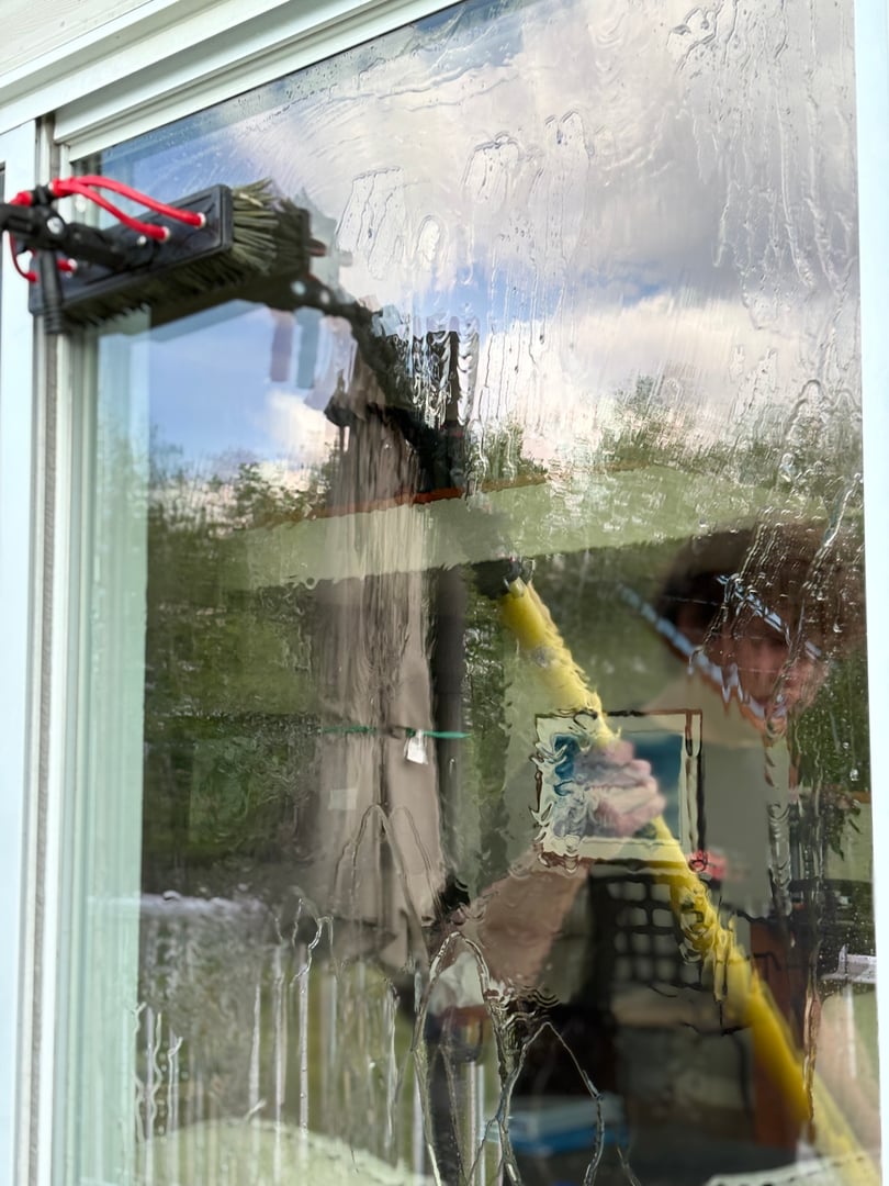 Person cleaning a large glass window with a long brush and water, visible through the wet, soapy surface.