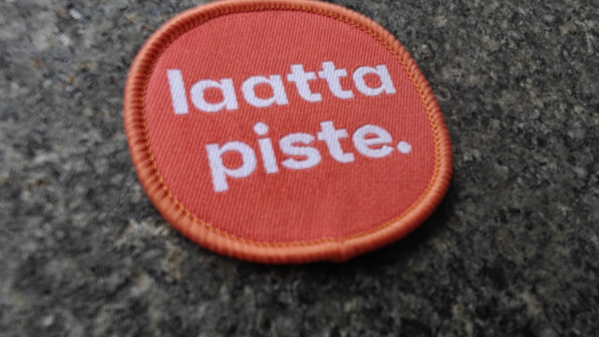 Orange circular patch with the words laattapiste in white, placed on a textured gray surface.