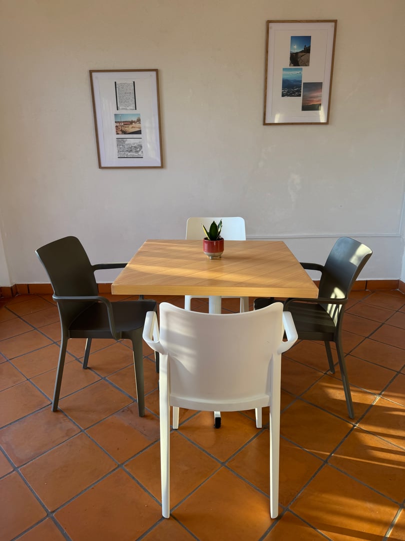 A square wooden table with a small plant in the center, surrounded by four chairs on a tiled floor, with framed photos on the wall.